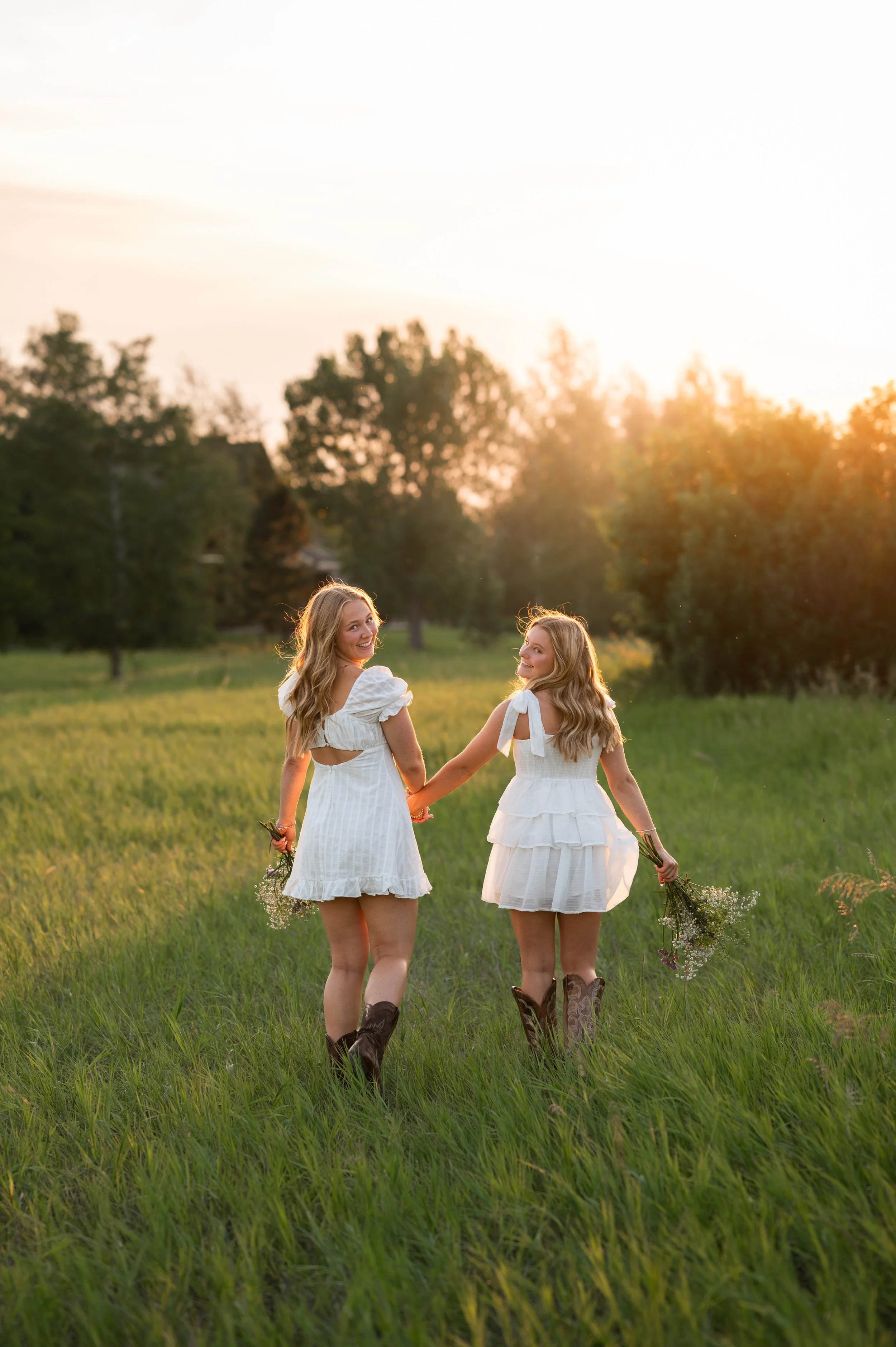 Two young women in white dresses holding flowers walk hand in hand through a grassy field at sunset.