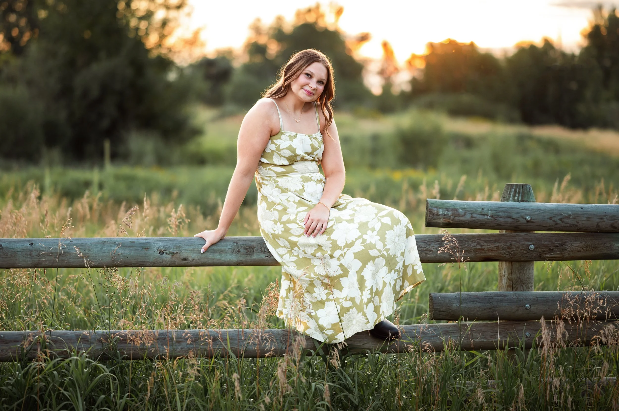 A young woman with long, wavy hair sitting on a wooden fence in a field at sunset, wearing a light green and white floral dress.