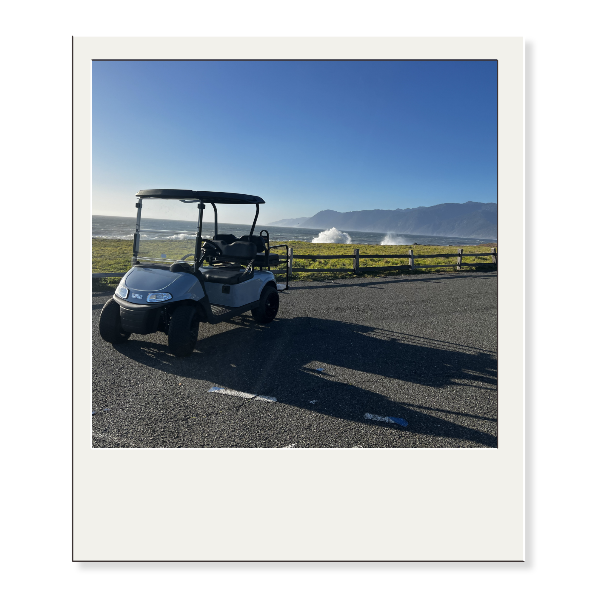 A grey and black golf cart parked on a paved road next to a grassy area by the ocean, with mountains in the background and waves crashing on the shore under a clear blue sky.