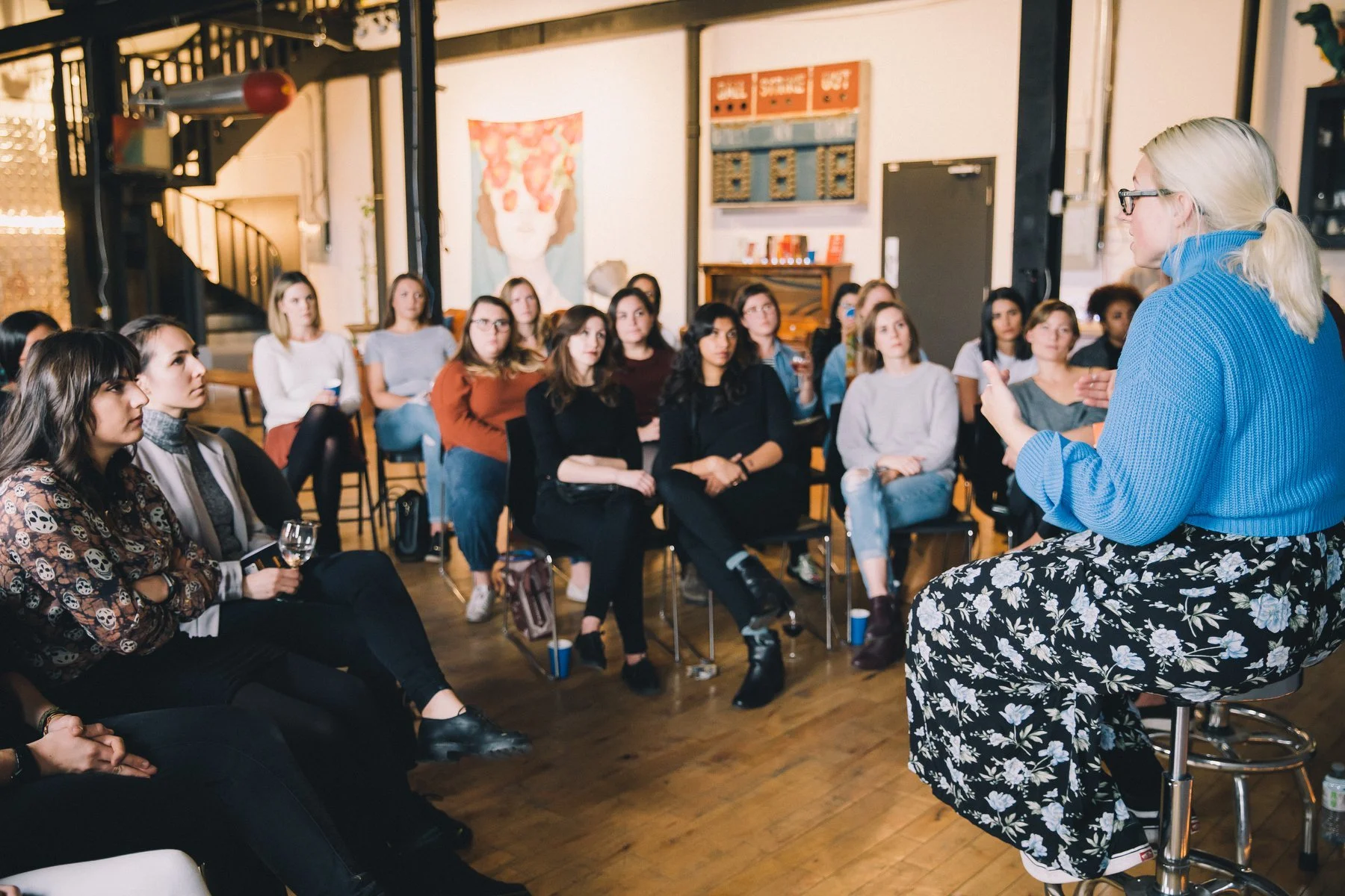 A woman in a blue sweater and floral skirt speaking to an audience of women in a room with artwork and wooden flooring.