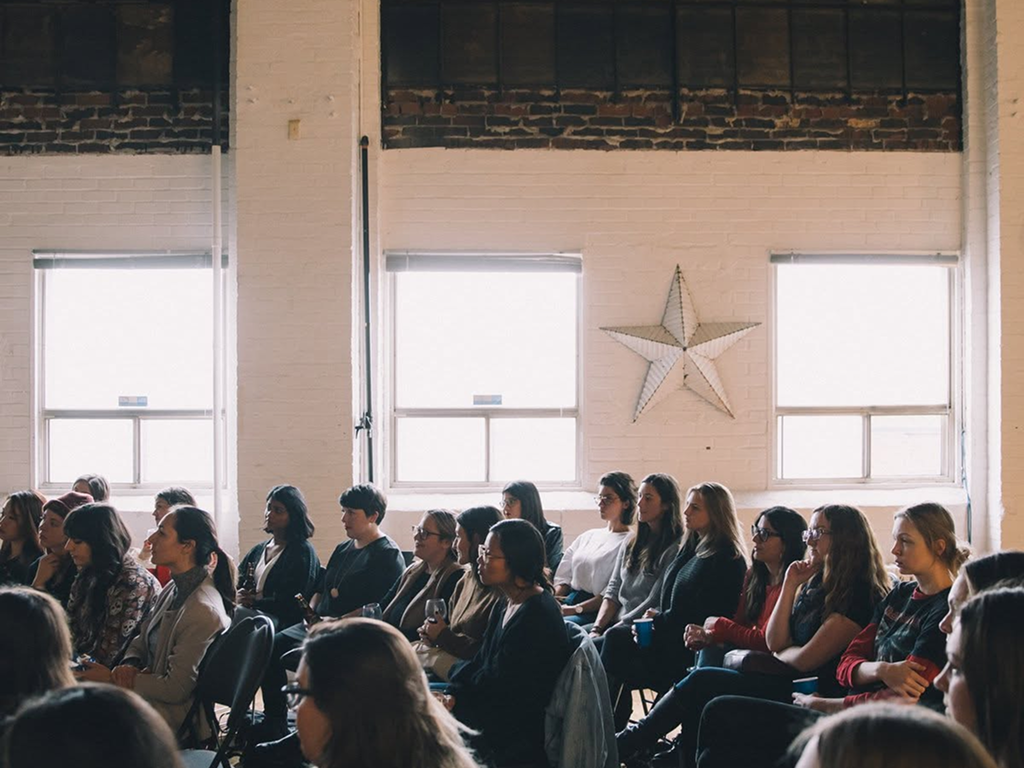A woman in a blue sweater and floral skirt speaking to an audience of women in a room with artwork and wooden flooring.