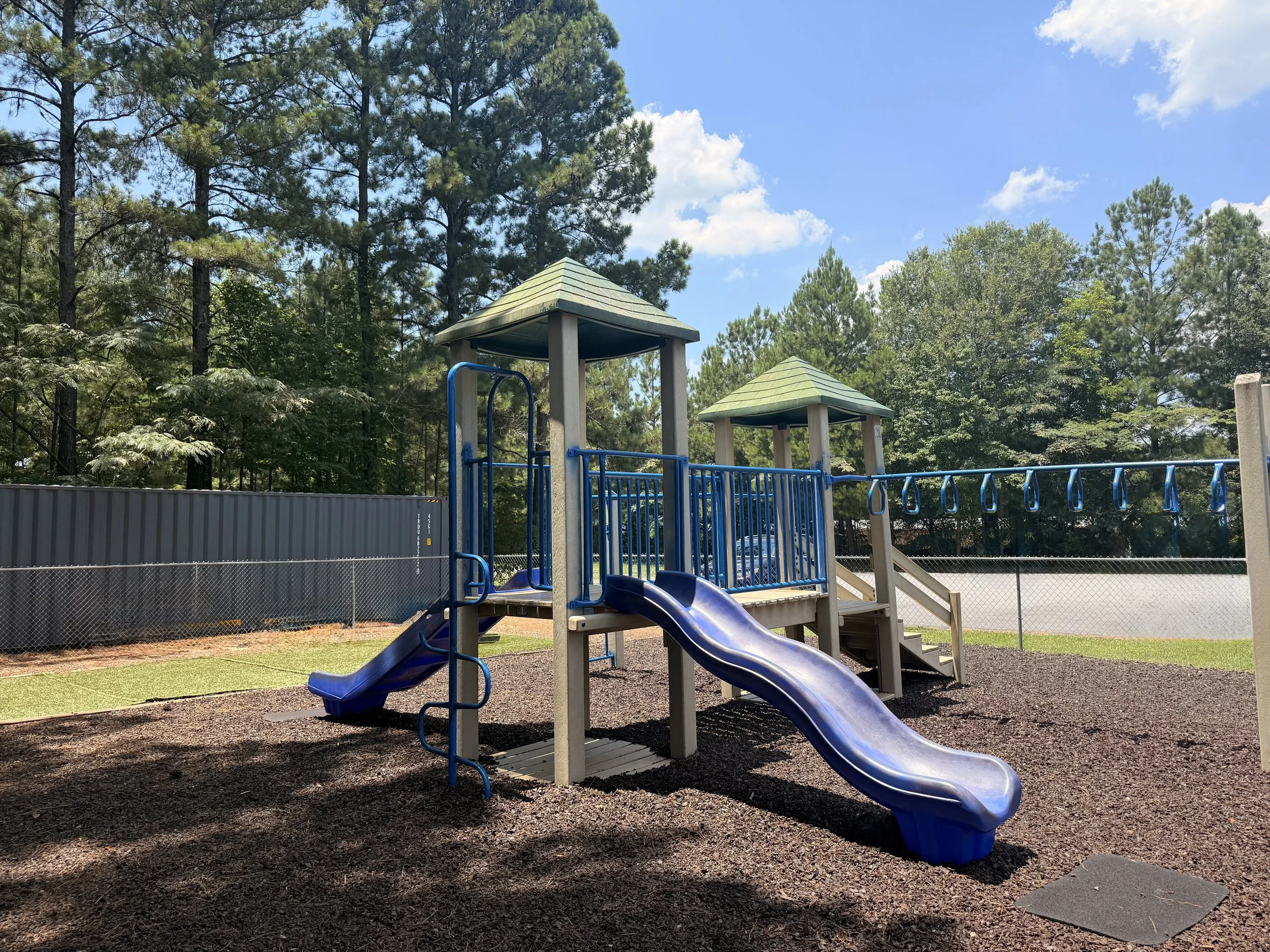 Children's playground with blue slides and climbing equipment, set on wood chips, surrounded by trees and a fence on a sunny day.