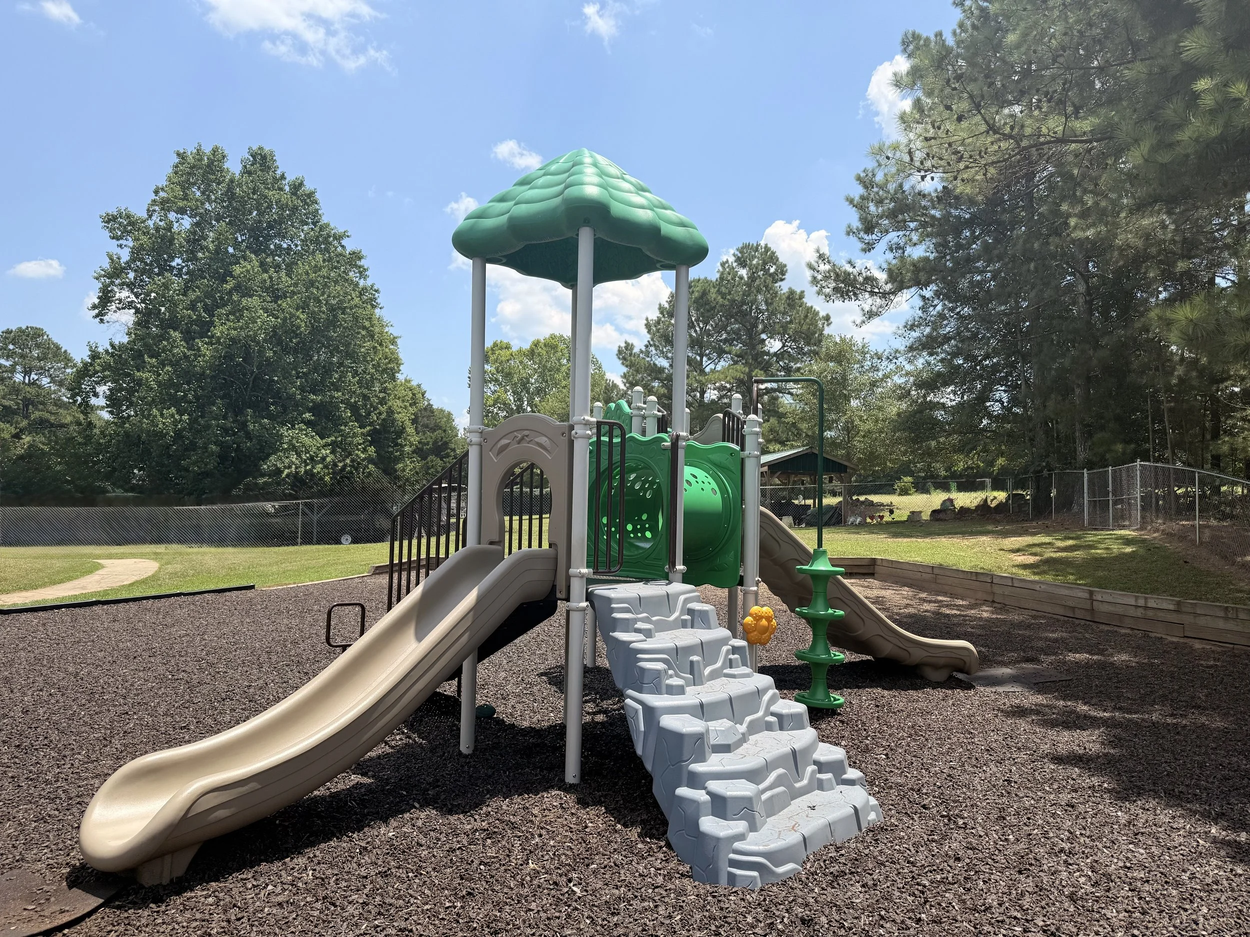 Colorful playground with slides, climbing structures, and a green canopy, surrounded by trees and a chain-link fence on a sunny day.