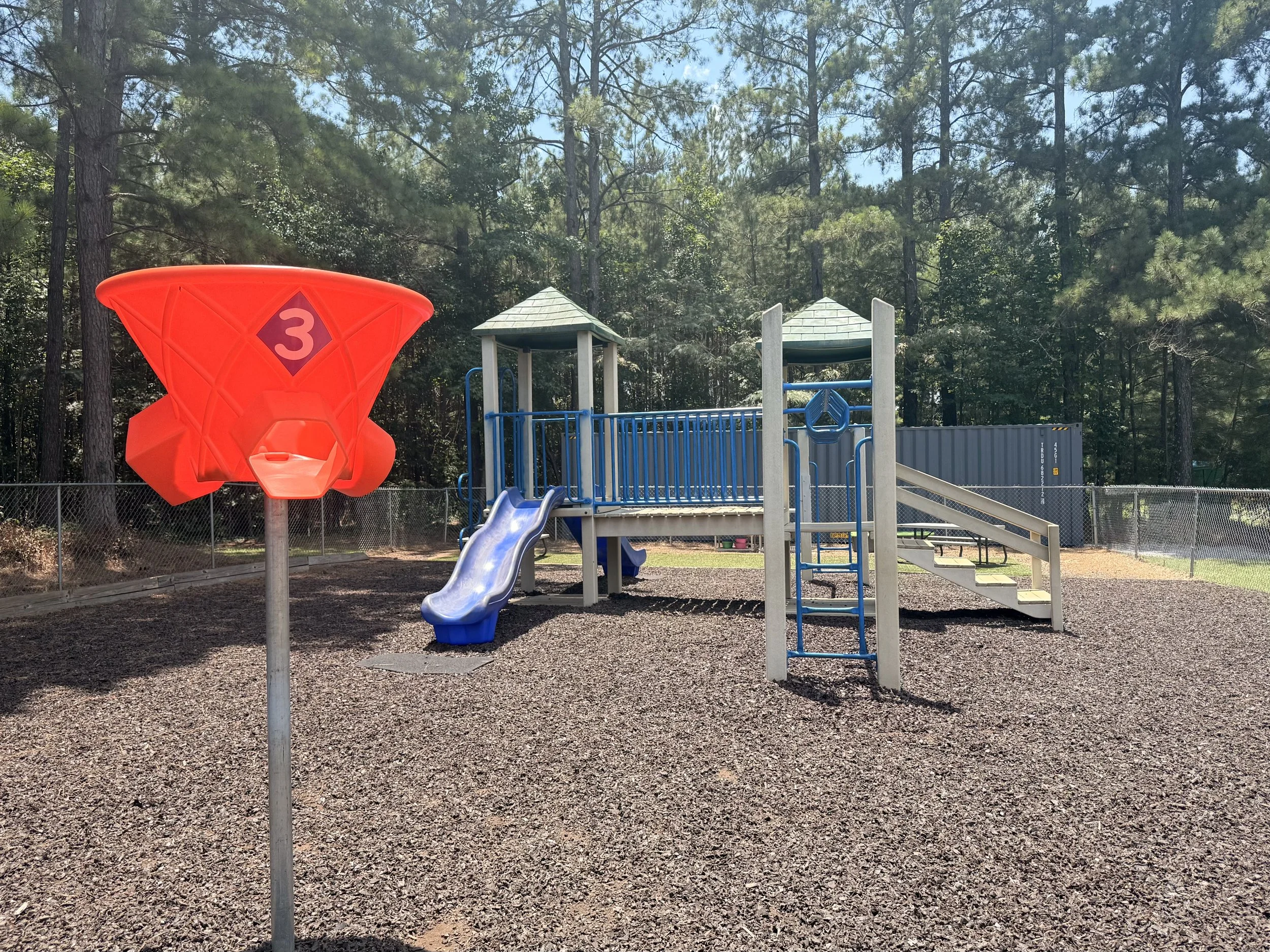 A playground with a red play structure shaped like a rocket with the number 3 on it, a slide, and a climbing ladder, surrounded by trees and a chain-link fence.