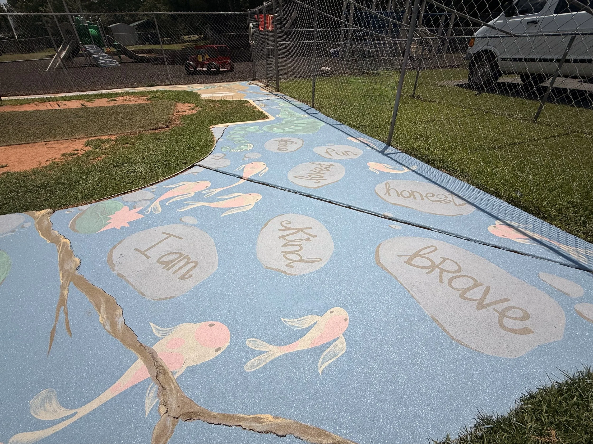 Painted sidewalk with playful fish and motivational words like "I am brave" and "kind" in a park area, with a playground and parked cars in the background.