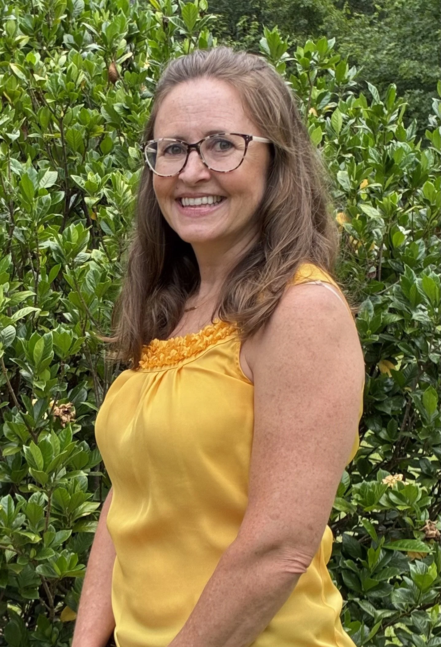 Smiling woman with long brown hair, glasses, and a yellow sleeveless top standing outdoors with green bushes in the background.