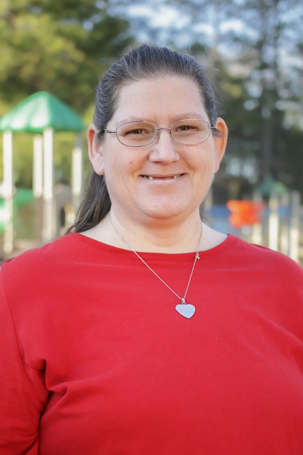 A woman with long dark hair, wearing glasses and a red top, smiling outdoors with trees and playground equipment in the blurred background.