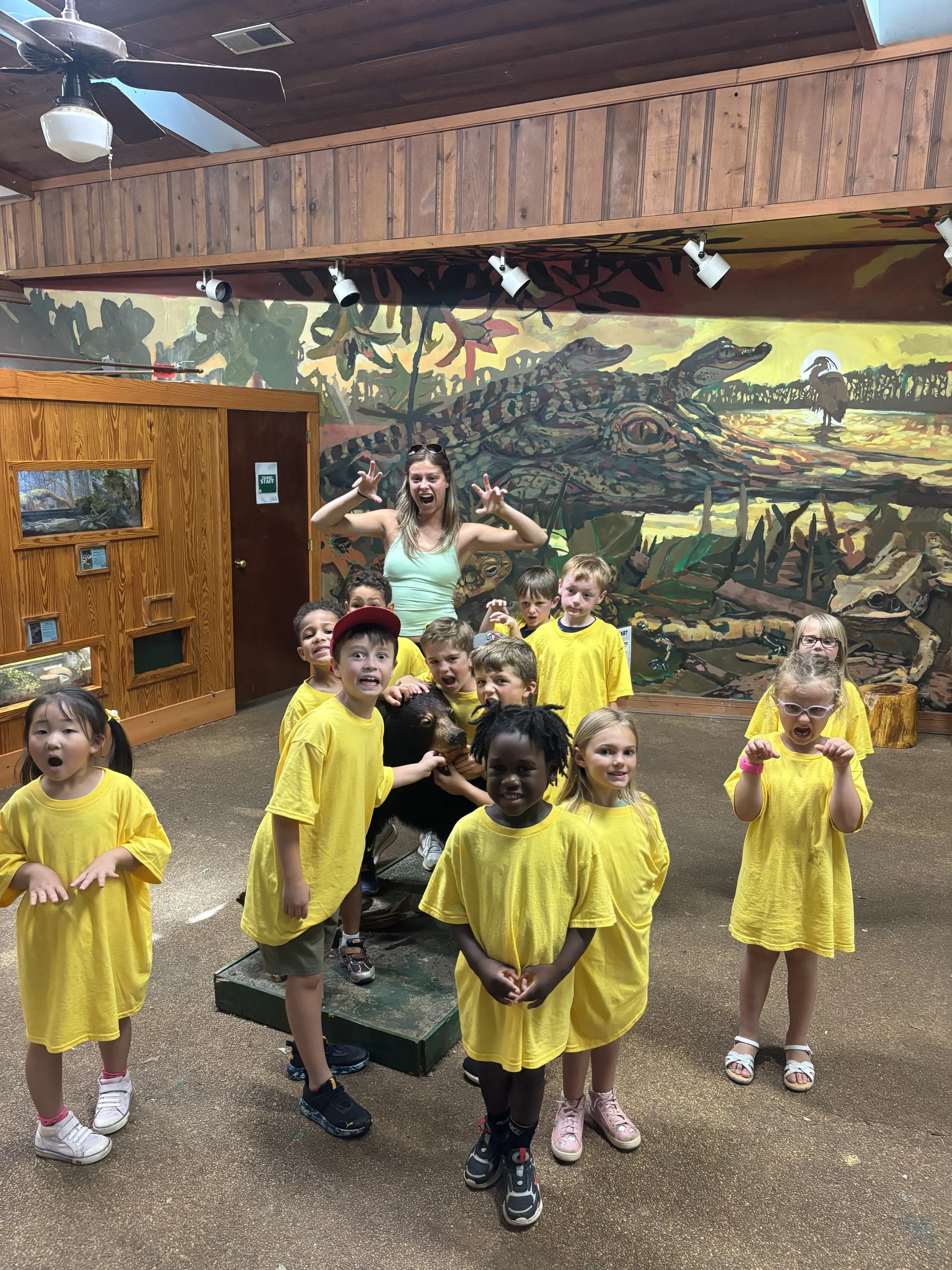 Group of children and a woman at a zoo exhibit with a crocodile mural in the background, children wearing yellow shirts, some with playful or surprised expressions, some pretending to be a crocodile. Masters Academy Oconee County Preschool Bogart, Wa