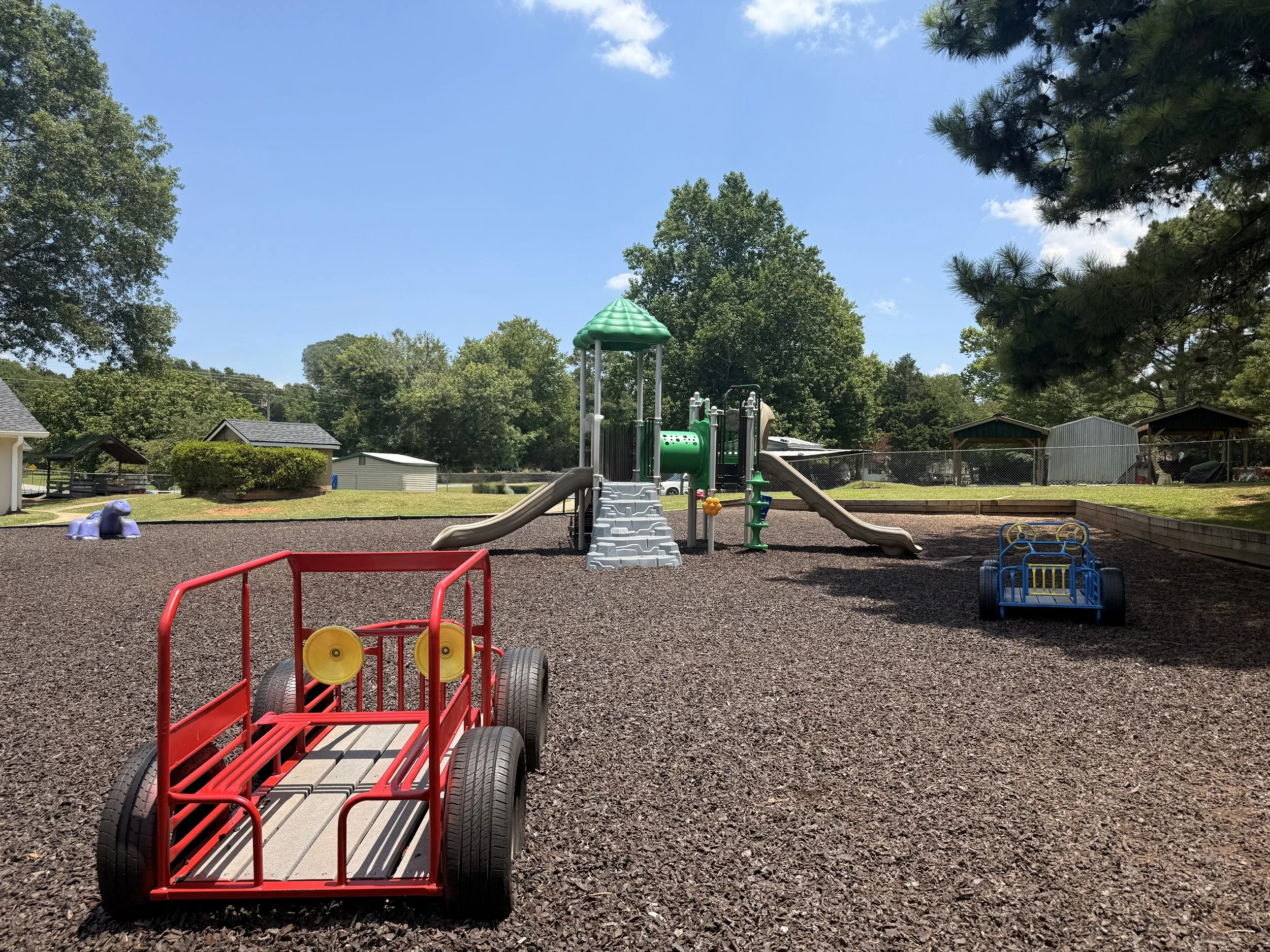 Empty playground with slides, a play car, a tunnel, and a purple isopod-shaped rocker, surrounded by trees and a clear blue sky.