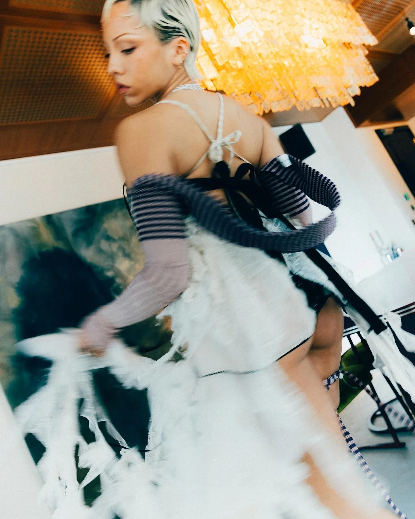 A woman in a black and white striped long-sleeve shirt with stripped arms and a white fluffy skirt poses indoors with warm lighting and artwork on the wall behind her.