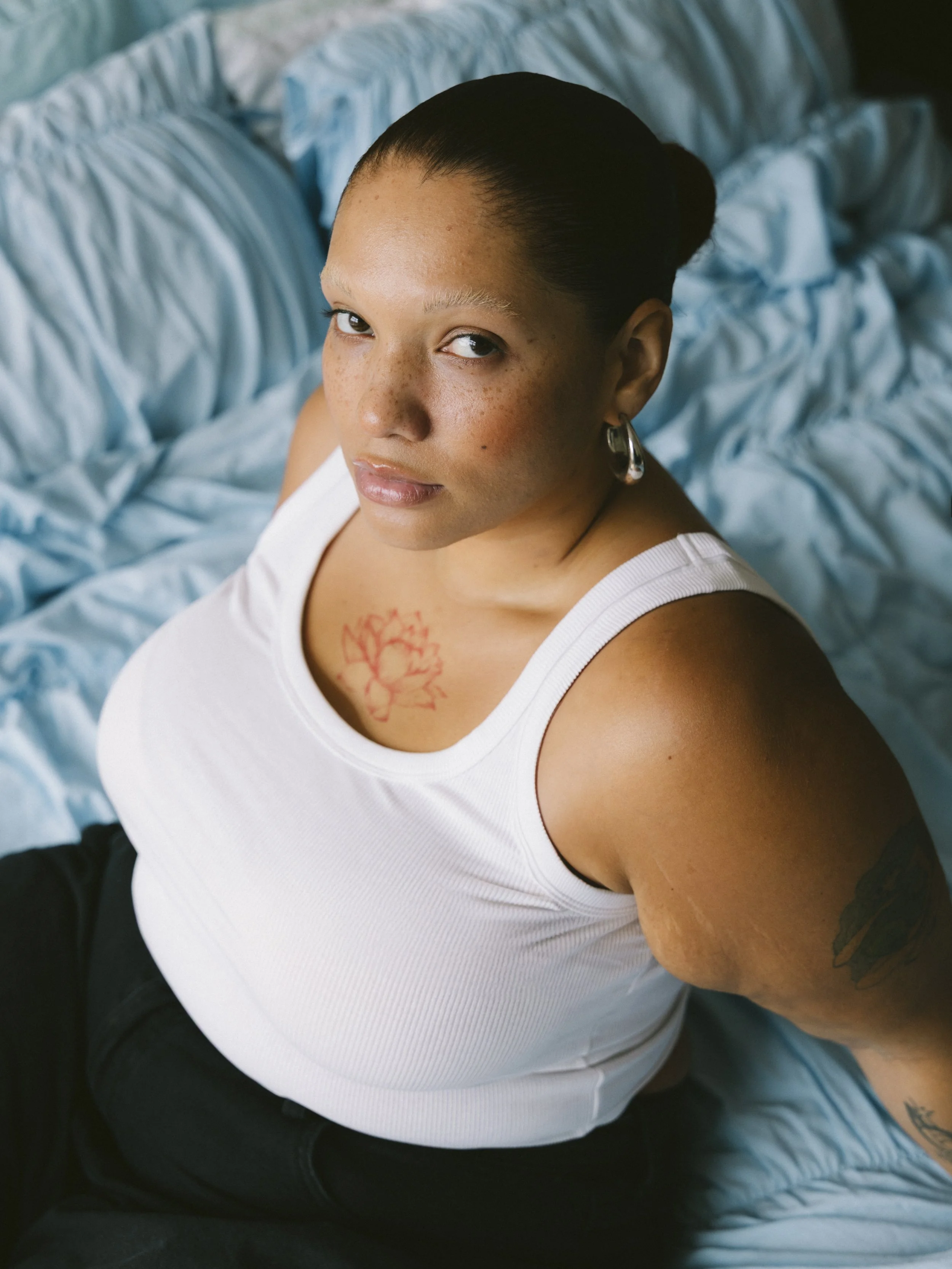 A woman with dark hair in a bun, wearing a white tank top, sitting on a bed with light blue bedding, looking up at the camera.