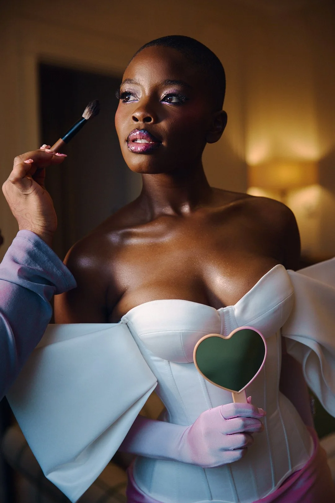 A woman with a shaved head applying makeup, holding a small mirror, wearing a white off-shoulder top with puffy sleeves, pink gloves, and a heart-shaped mirror, in a warmly lit room.