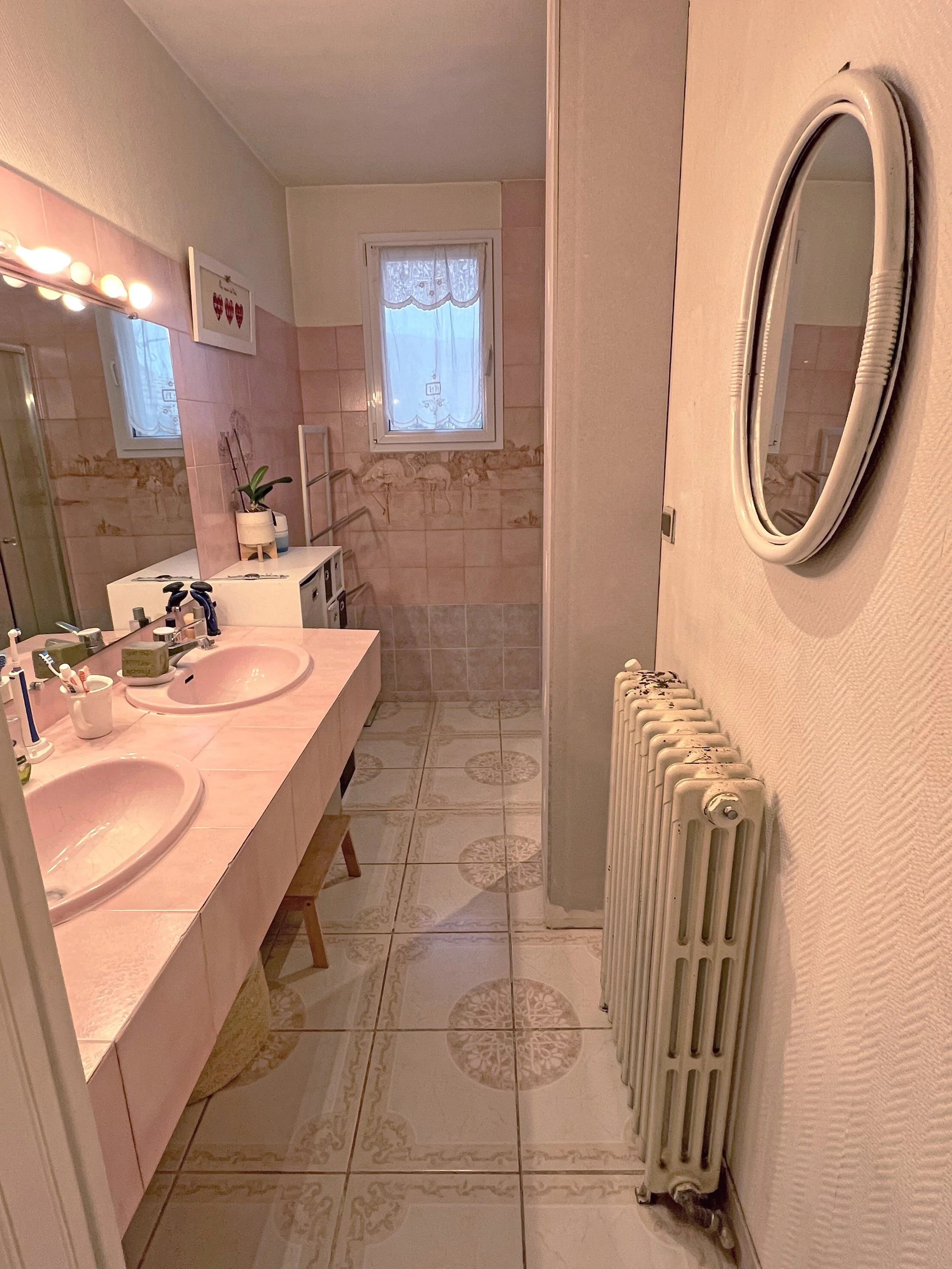 Salle de bain avec un double lavabo rose, un miroir rond, une fenêtre avec rideau en dentelle, un radiateur blanc et un miroir rectangulaire, décorée dans un style vintage.