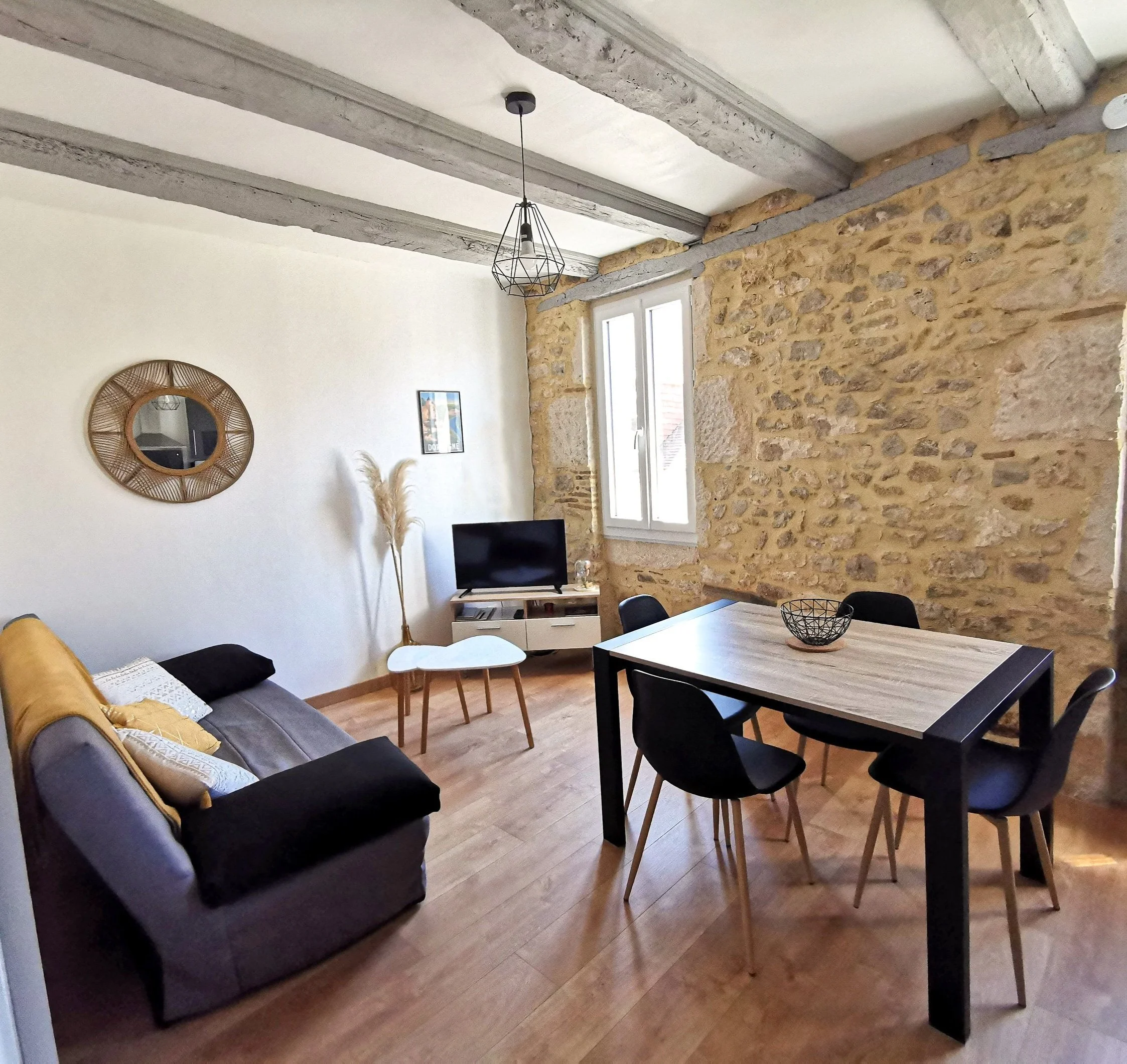 Salon avec canapé gris, table à manger en bois et chaises noires, mur en pierre apparente, fenêtre, télévision, miroir rond en rotin, décor avec des plantes et une lampe suspendue.