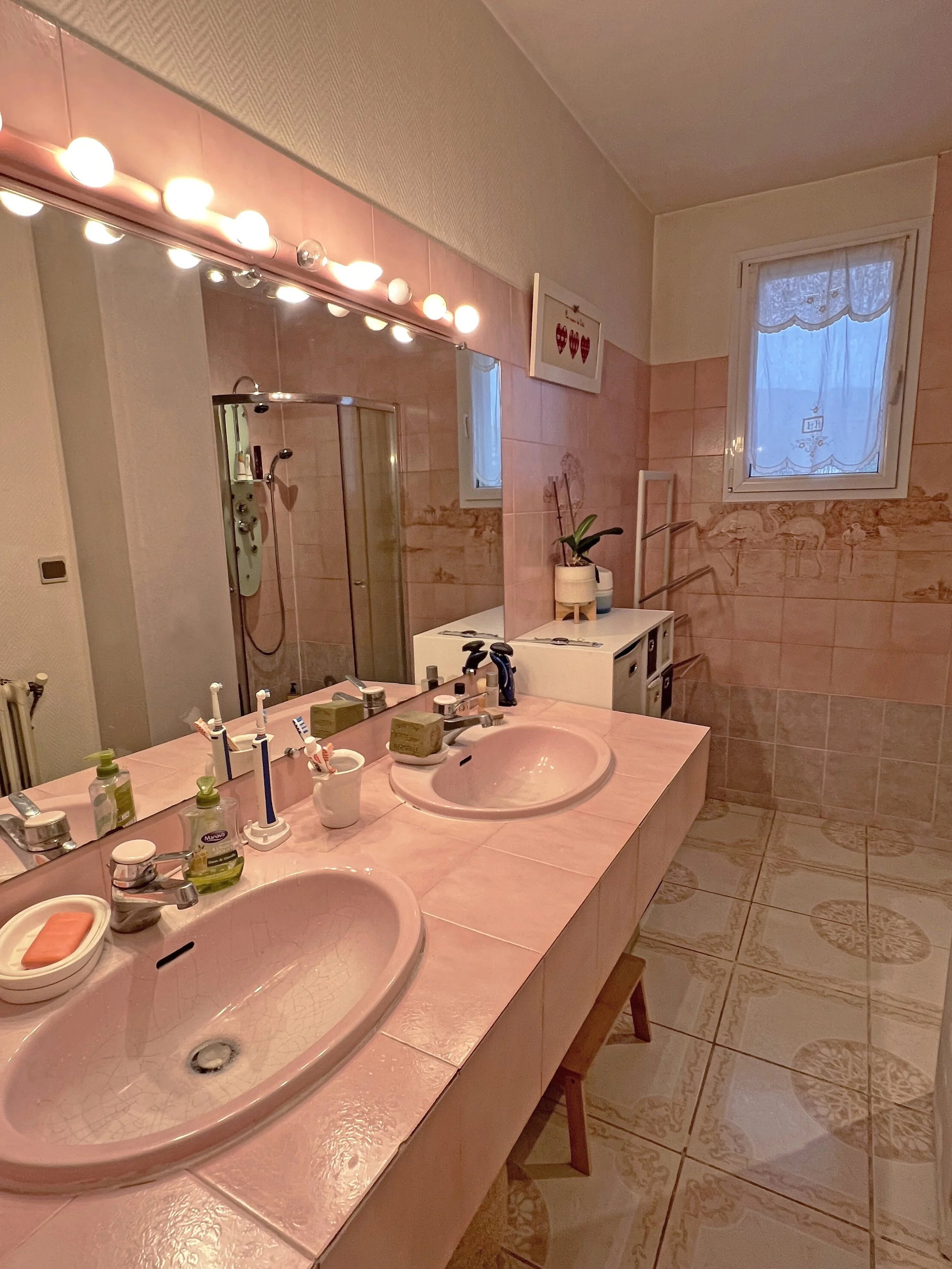 Salle de bain avec double lavabo rose, grande baignoire, miroir, éclairage à ampoules, fenetre avec voilage, décoration florale.