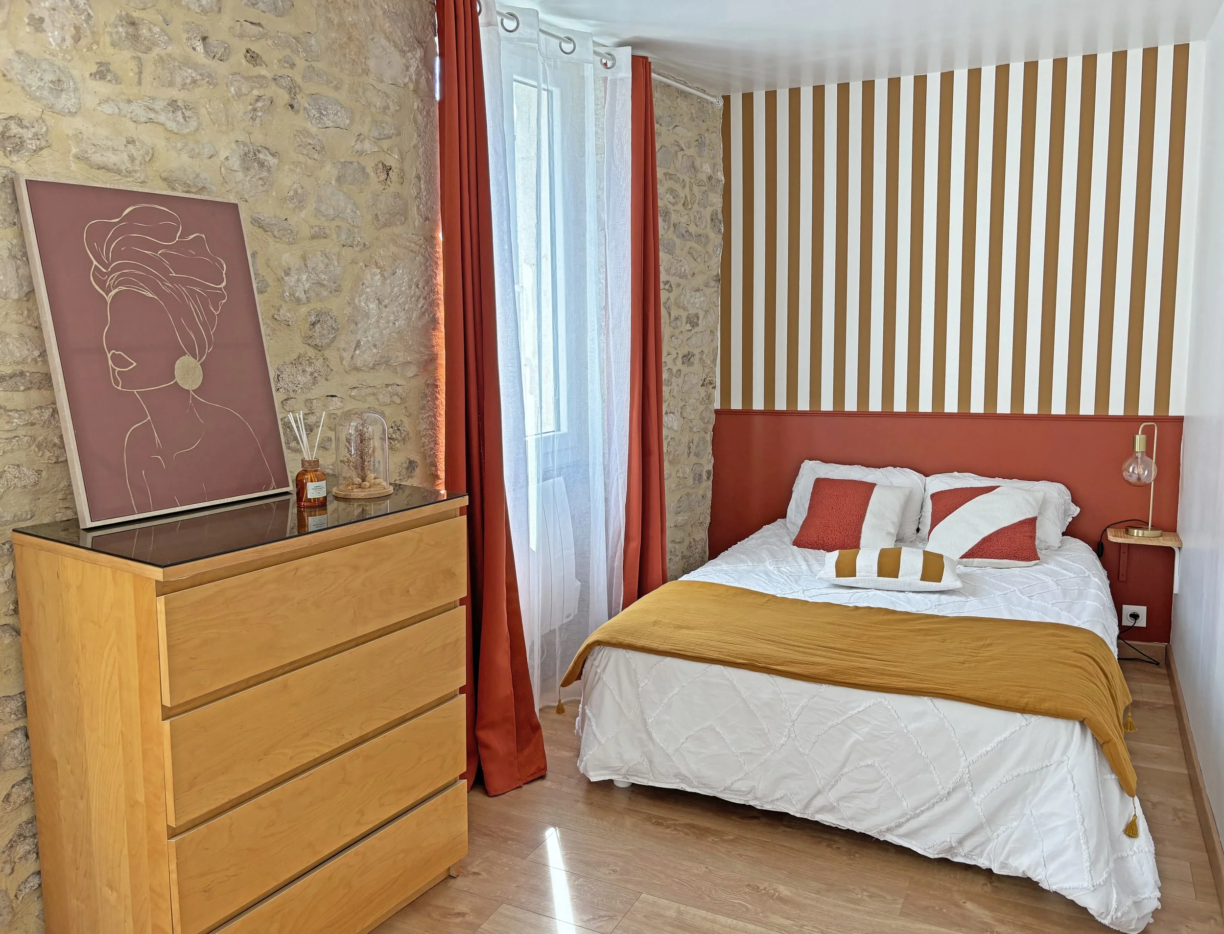 Chambre avec mur en pierre, lit avec oreillers blancs et decors rouges et or, une commode en bois, tableau d'une femme avec un turban sur la tête, rideaux rouges et blancs, et un mur avec rayures jaunes et blanches.