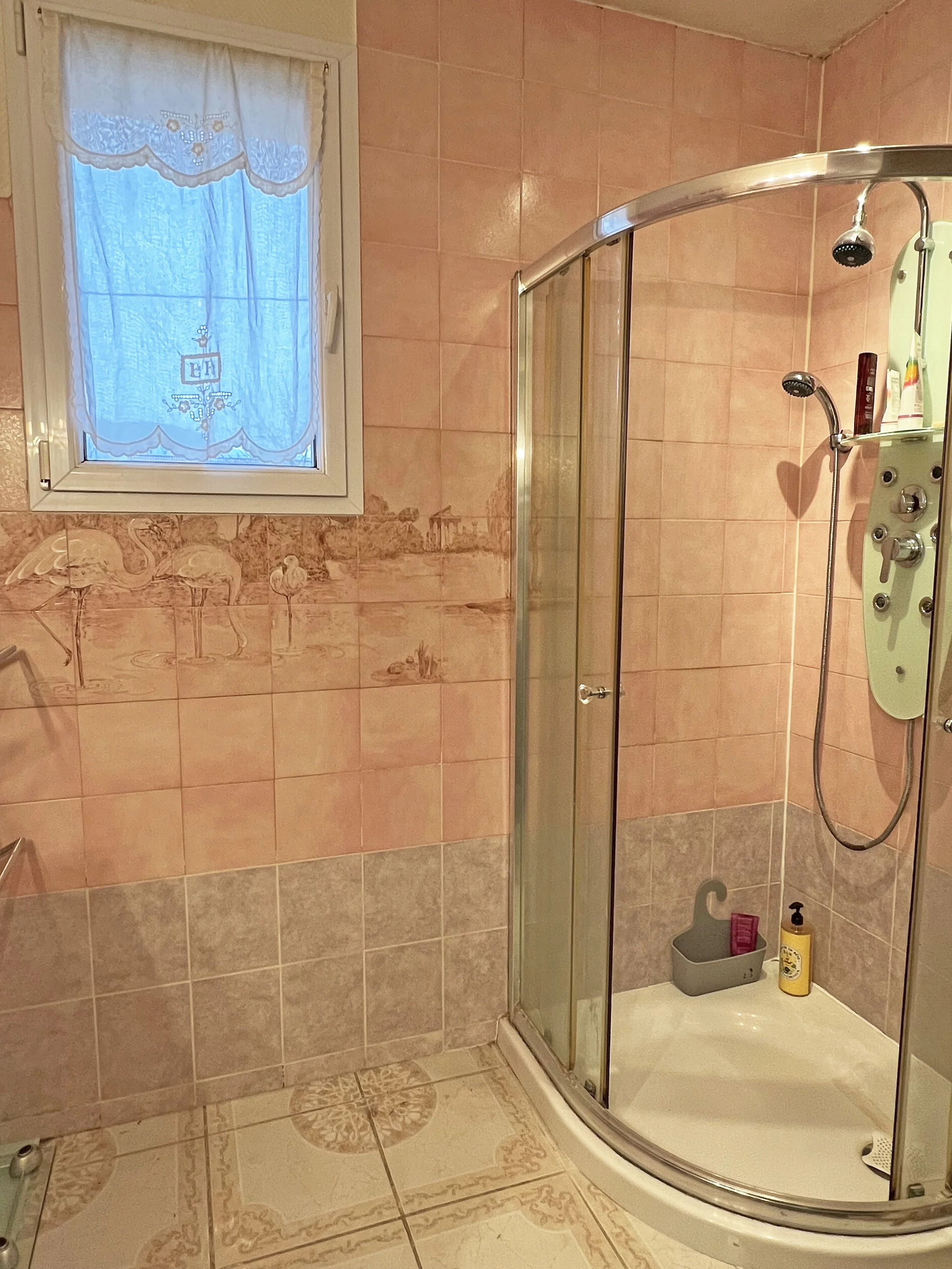 Salle de bain avec douche à l'italienne, mur en carrelage rose, fenêtre à voilage, sols en céramique avec mosaïque, étagère à produits de bain.