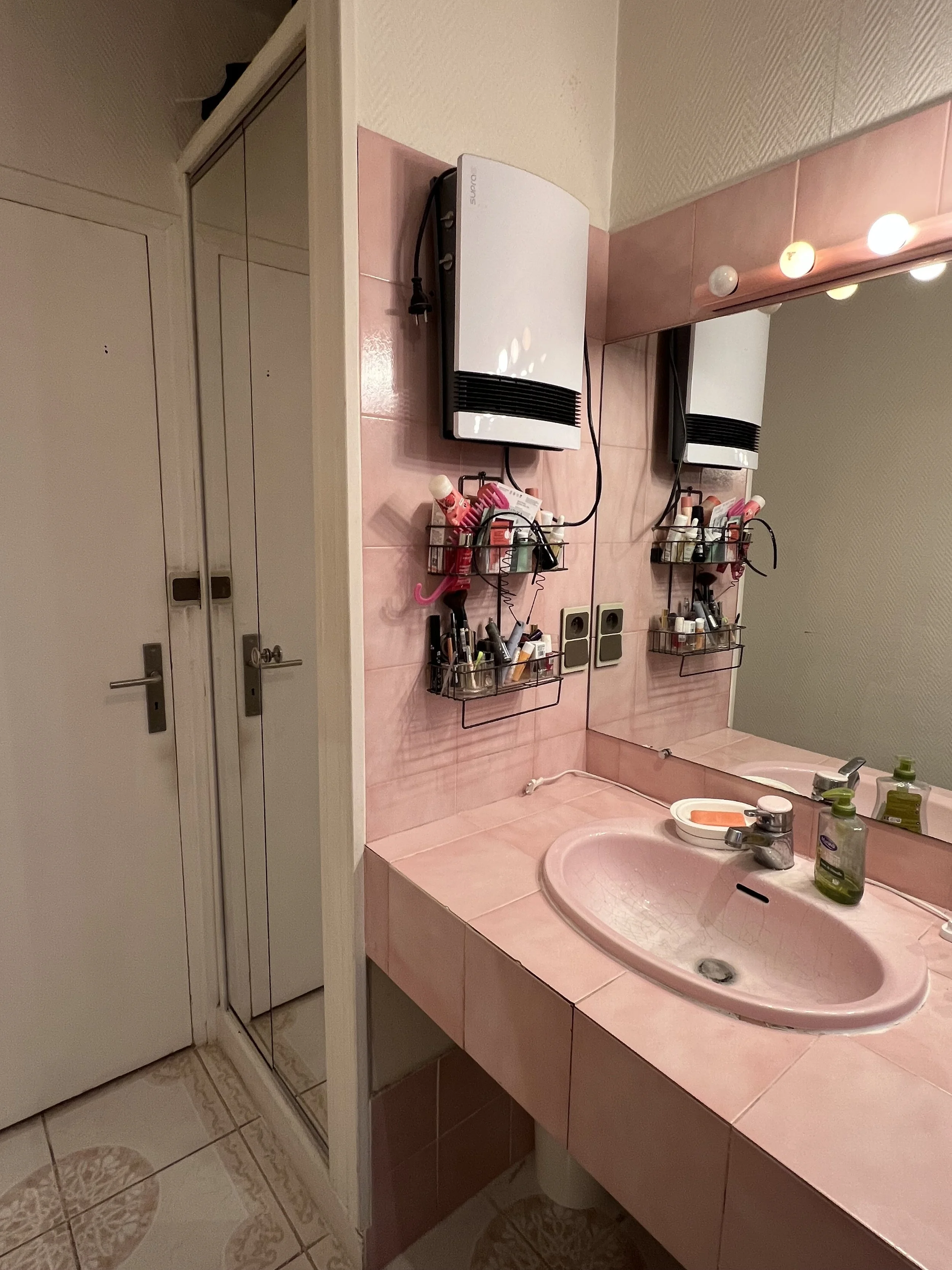 Salle de bain avec un miroir, un lavabo rose, des produits de toilette, un sèche-serviettes électrique, et une porte de cabine de douche.