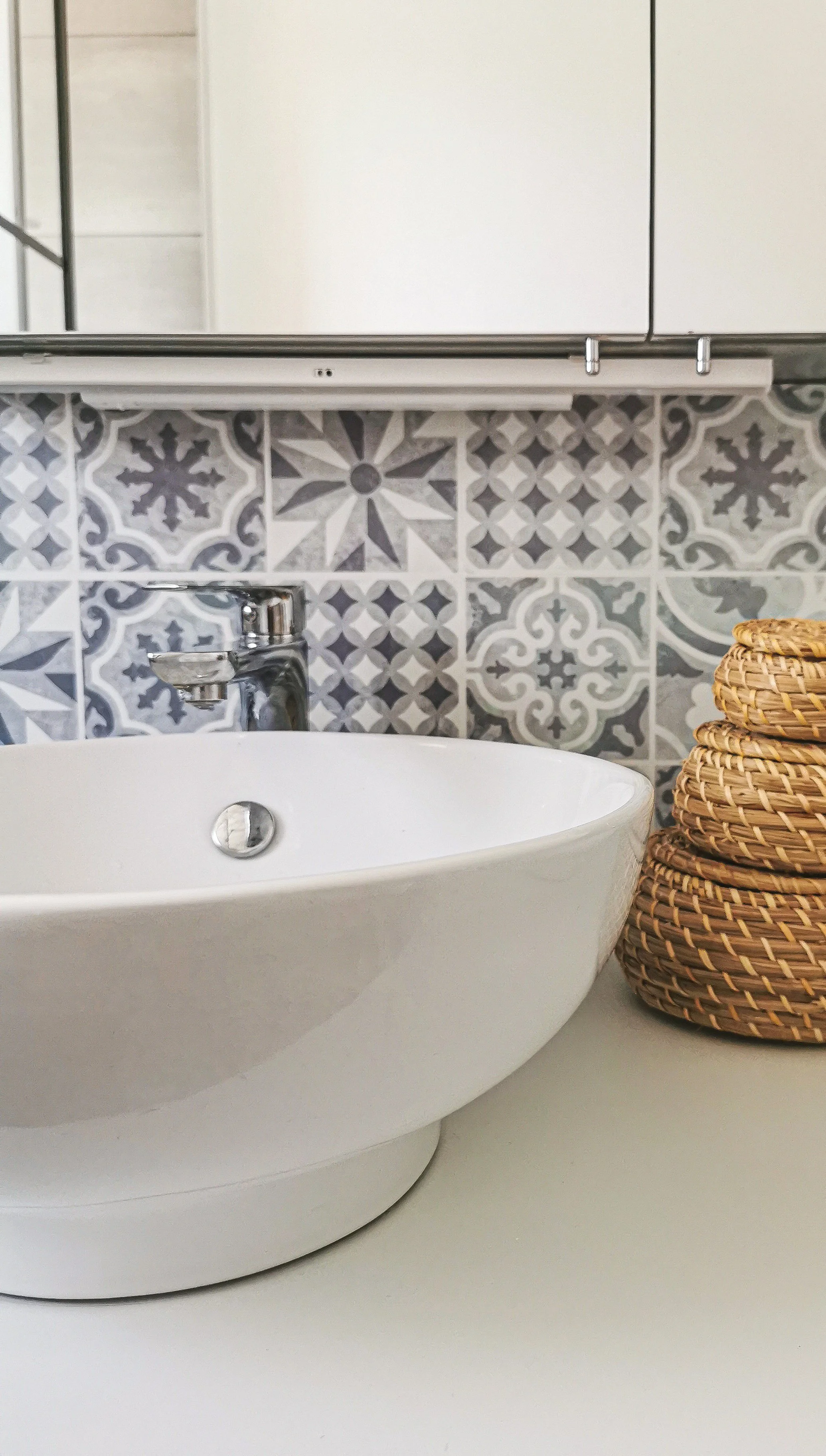 Lavabo blanc rond sous un miroir, avec un robinet en métal et un panier en rotin à côté, en arrière-plan une cloison avec des carreaux en céramique gris et blanc à motifs géométriques.