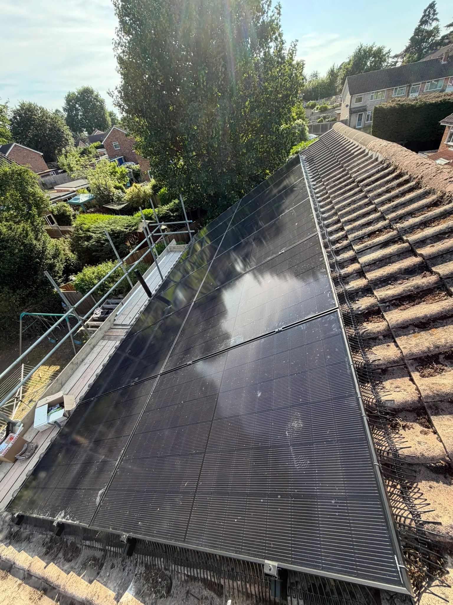 Solar panels installed on the sloped roof of a house, with surrounding trees and neighboring houses visible in the background.