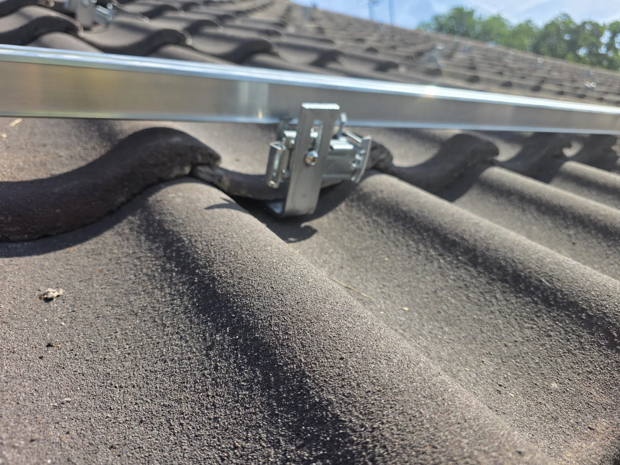 Close-up of a metal roof mounting bracket attached to a roof with dark shingles, with a metal rail secured to it, and a forest visible in the background.
