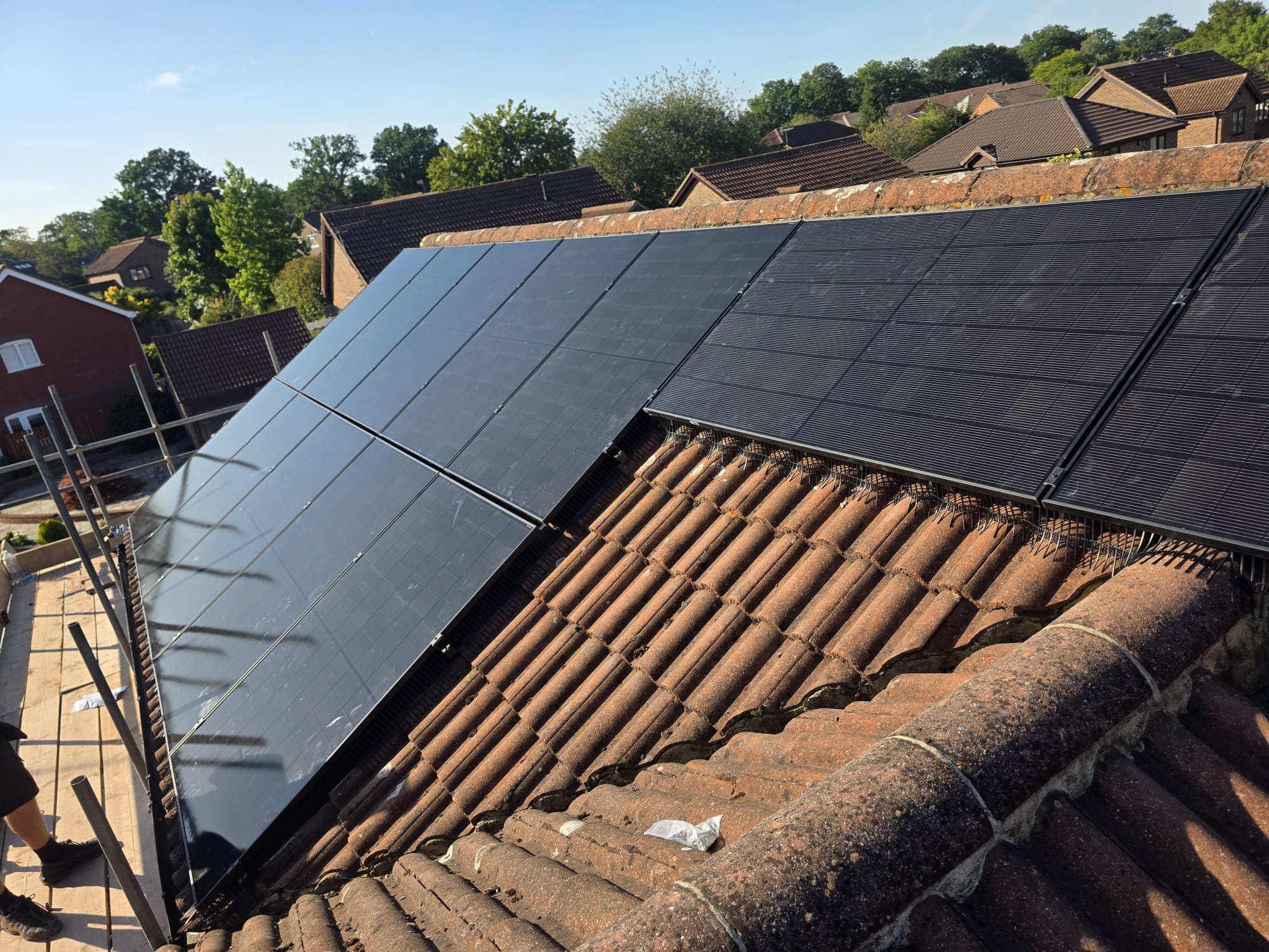 Solar panels installed on a sloped roof of a residential house, with neighboring houses and green trees in the background under a clear blue sky.
