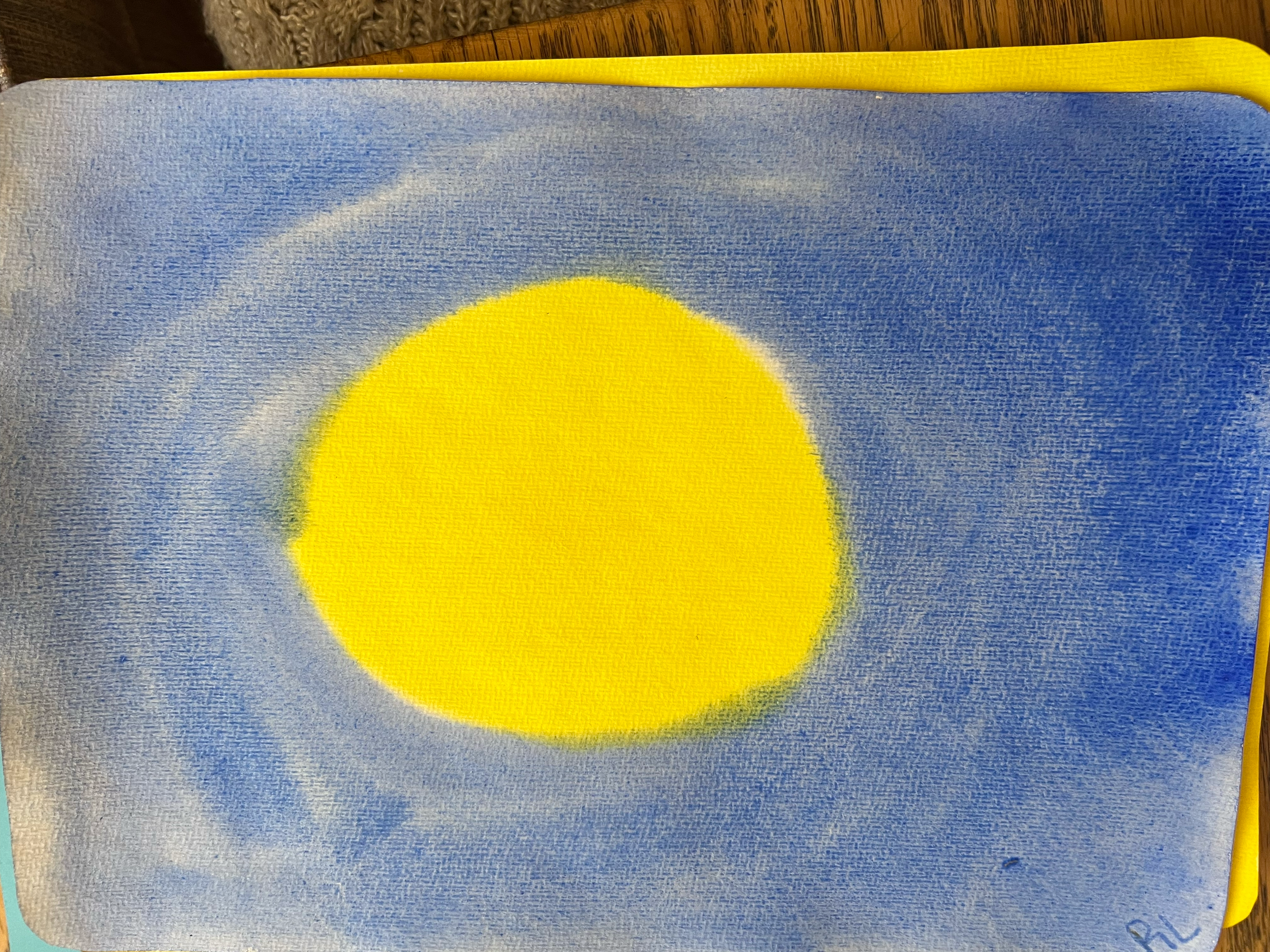 A drawing of a yellow sun with a blue sky background on a piece of paper.