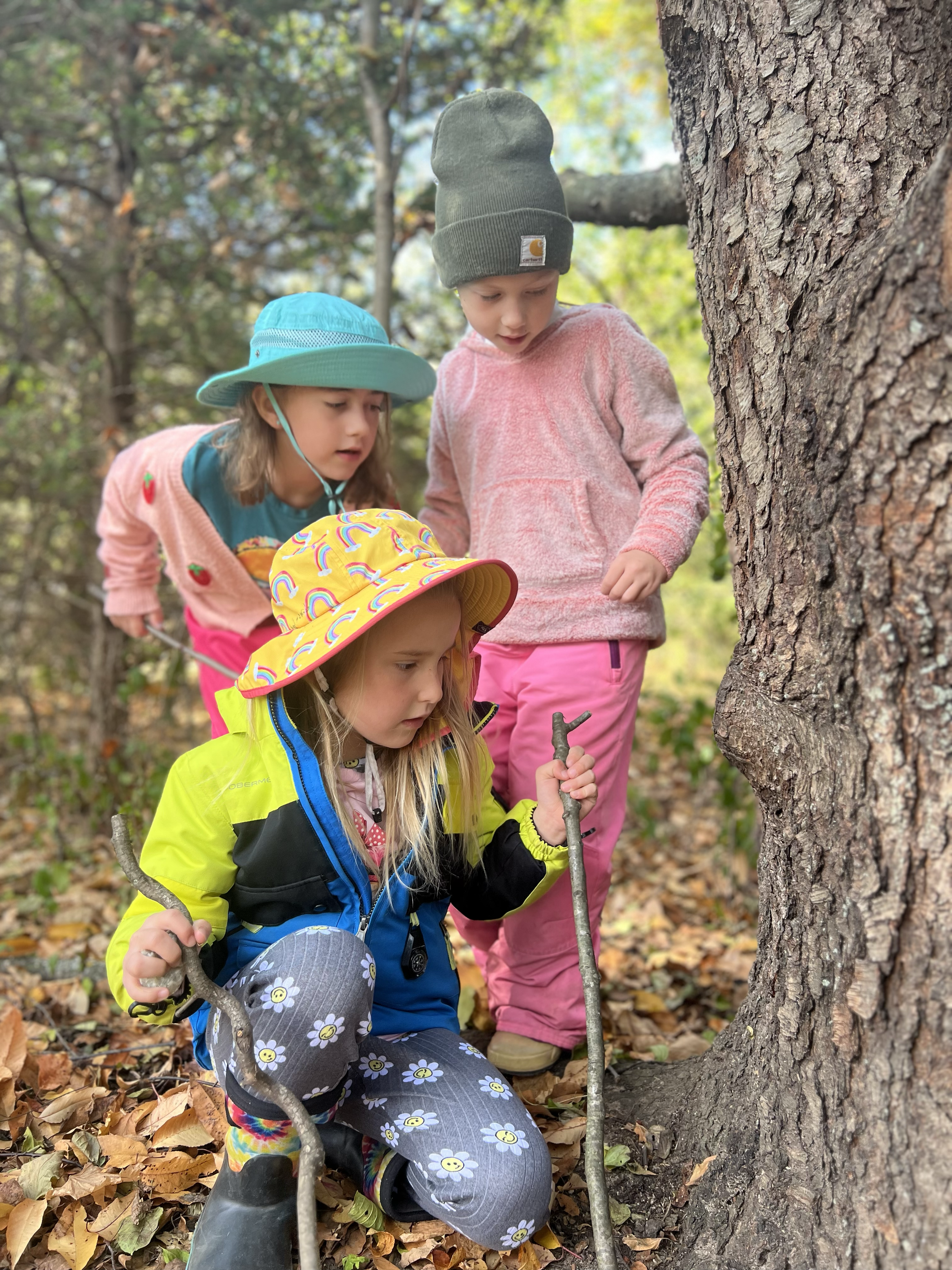 Three young children outdoors in a wooded area, closely inspecting the ground near a tree. One girl in a yellow rain jacket and a colorful rain hat is kneeling and holding a walking stick, while the other girl, in pink pants and a hoodie, and another