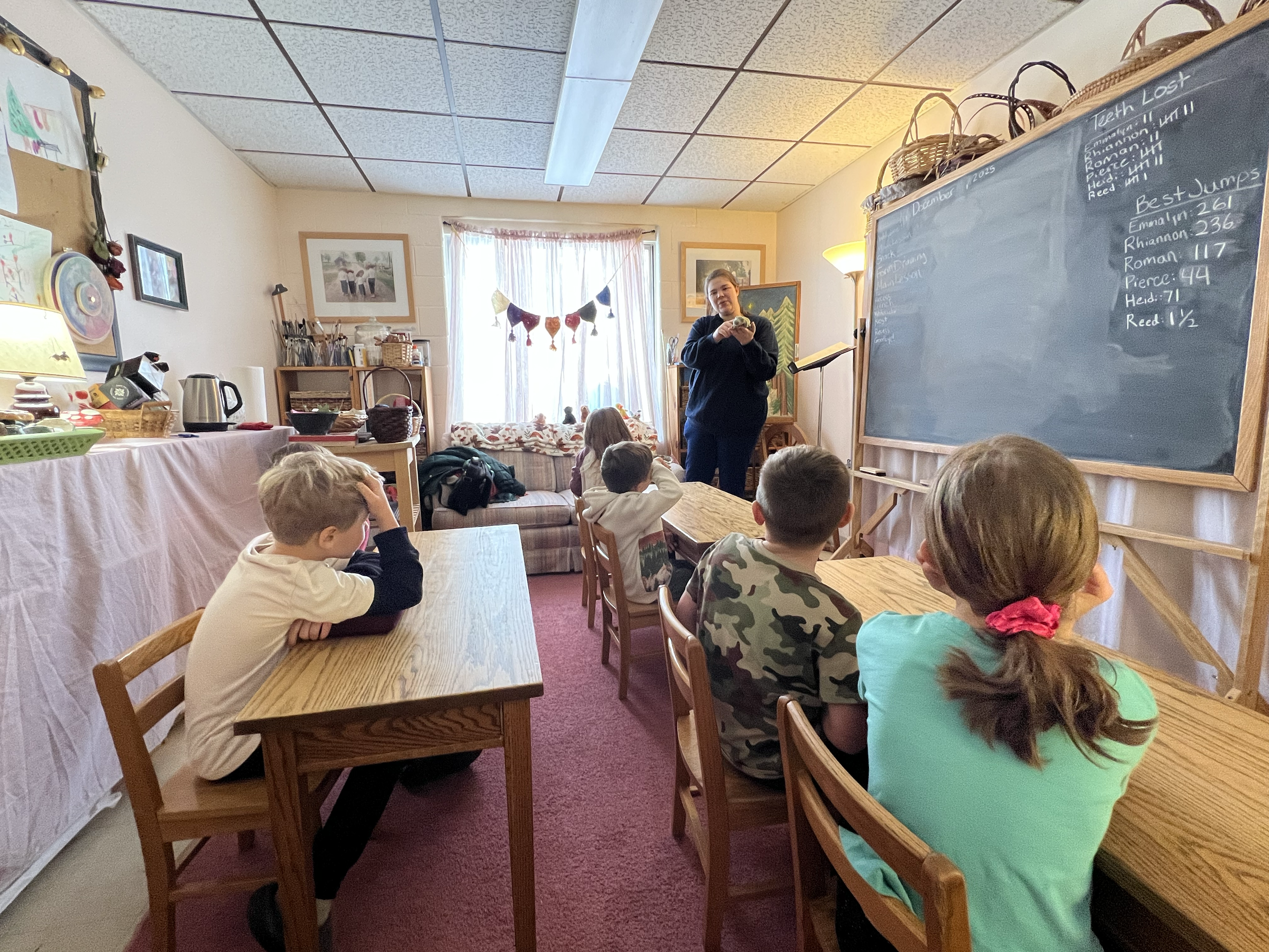 A classroom with five children sitting at desks facing a teacher in front of a chalkboard. The teacher appears to be holding a blanket. The classroom has a window with curtains, a couch, and various decorations and supplies on shelves.