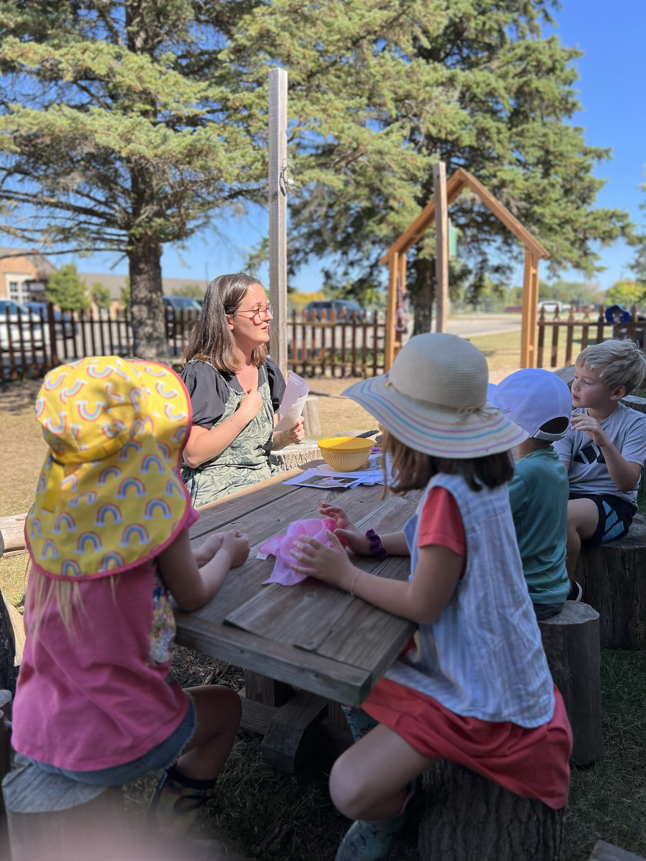 A woman with glasses reads to a group of children sitting at a rustic wooden table outdoors on a sunny day, with trees and a fence in the background.