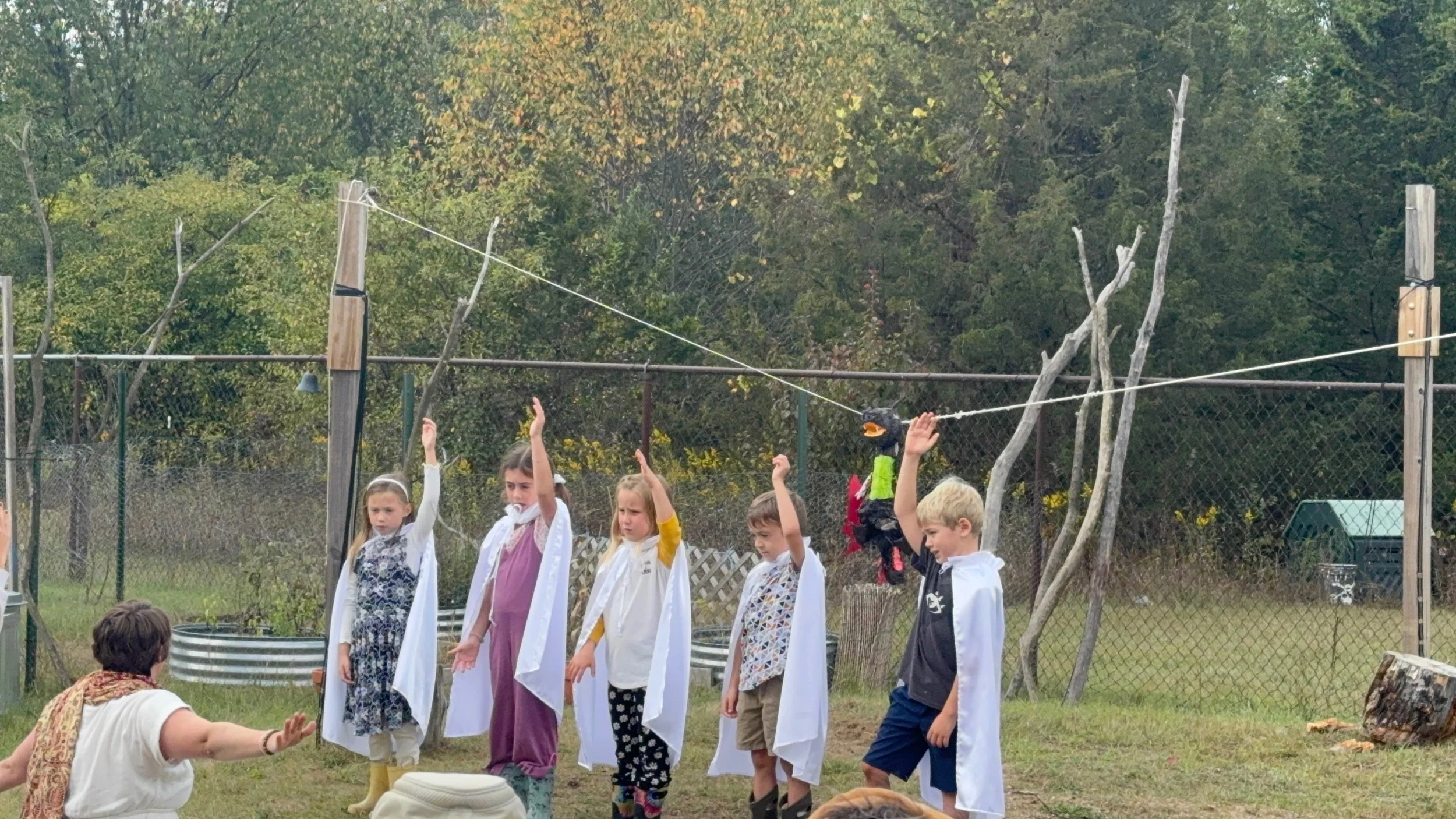 Children participating in a school play or performance outdoors, with some children raising their hands and one woman sitting in front of them.