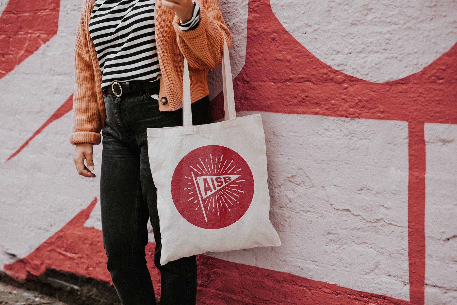 Person wearing an orange sweater and striped shirt holding a tote bag with "AISB" logo, standing against a red and white brick wall.