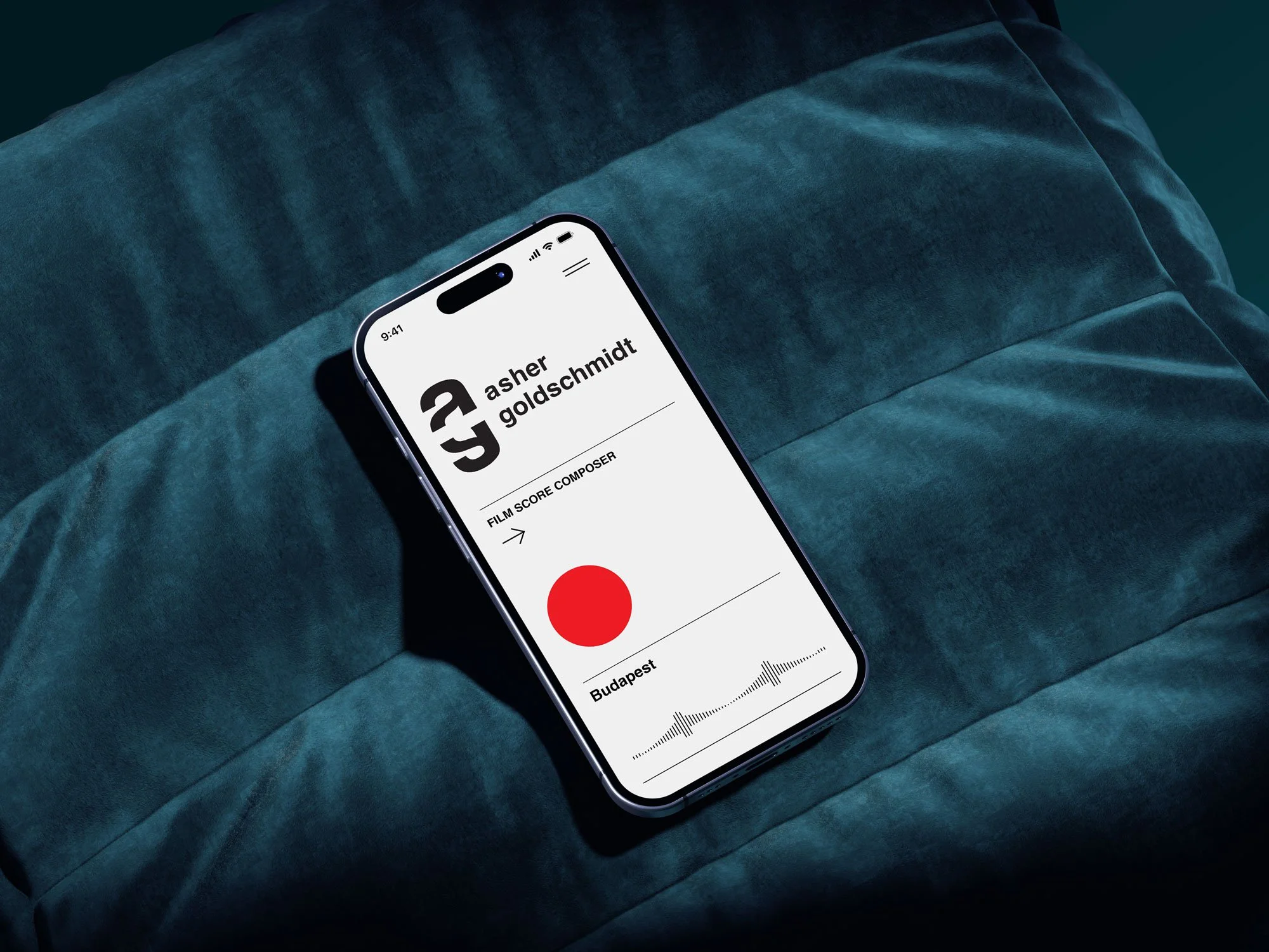 Smartphone displaying a voice recording app with a red record button, music waveform, and the name Asher Goldschmidt on a dark green velvet couch.