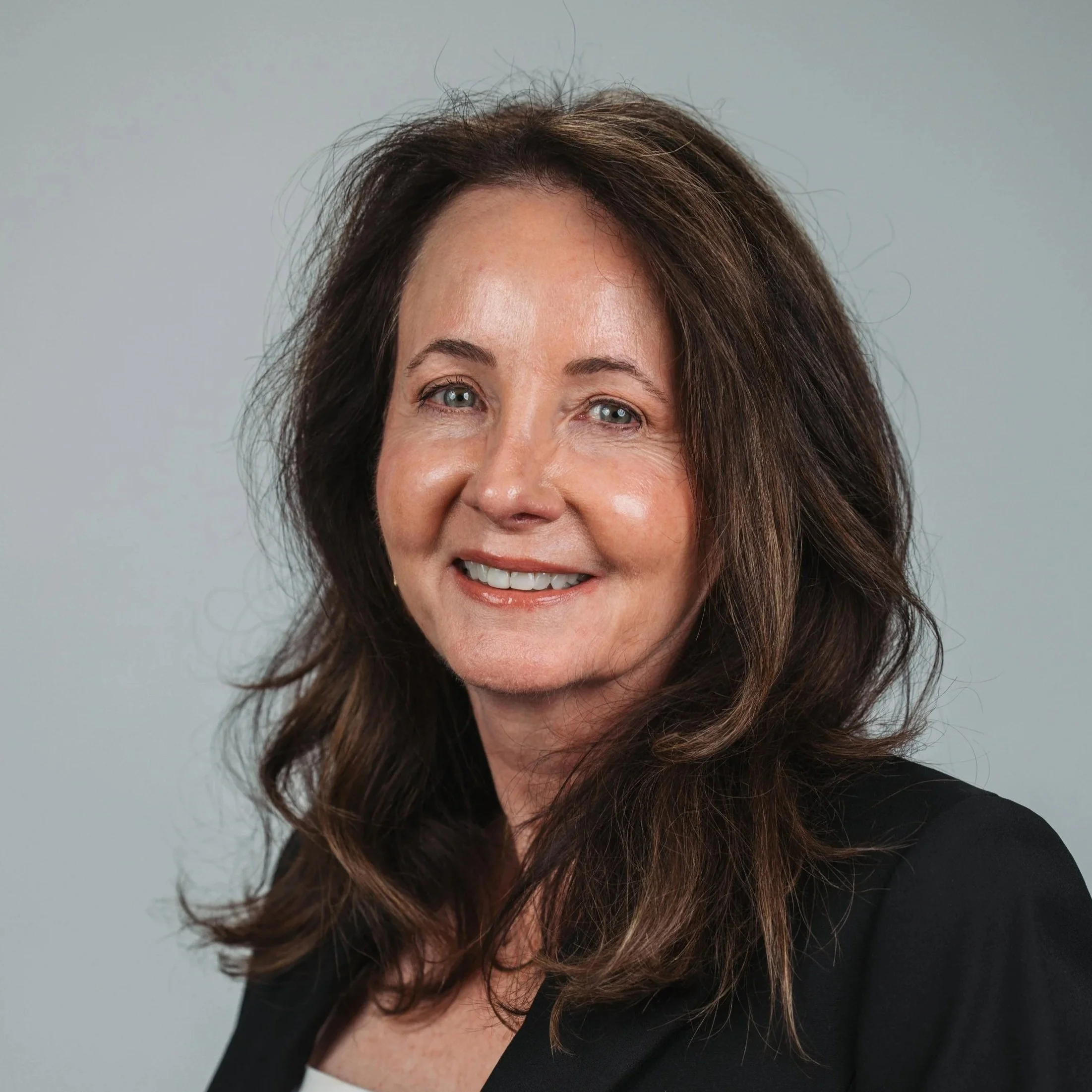 Dawn Soper, interim board chairman, with long, wavy brown hair and light skin, smiling and wearing a plaid blazer over a navy blue blouse, standing against a plain gray background.