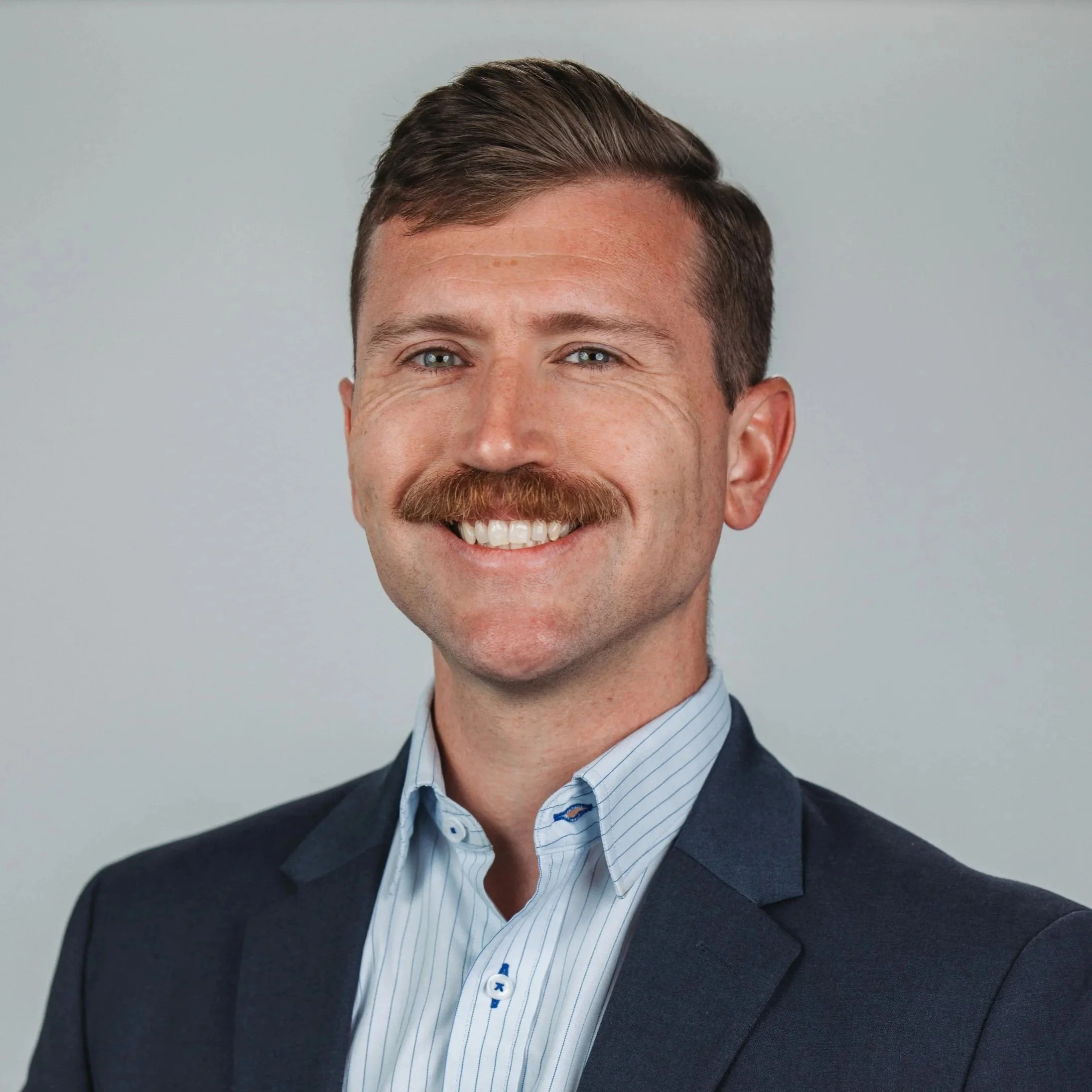 Brandon Hibbard, vice-chair and treasurer, with short brown hair and a beard, wearing a navy checkered blazer over a white shirt, smiling with arms crossed against a plain light gray background.