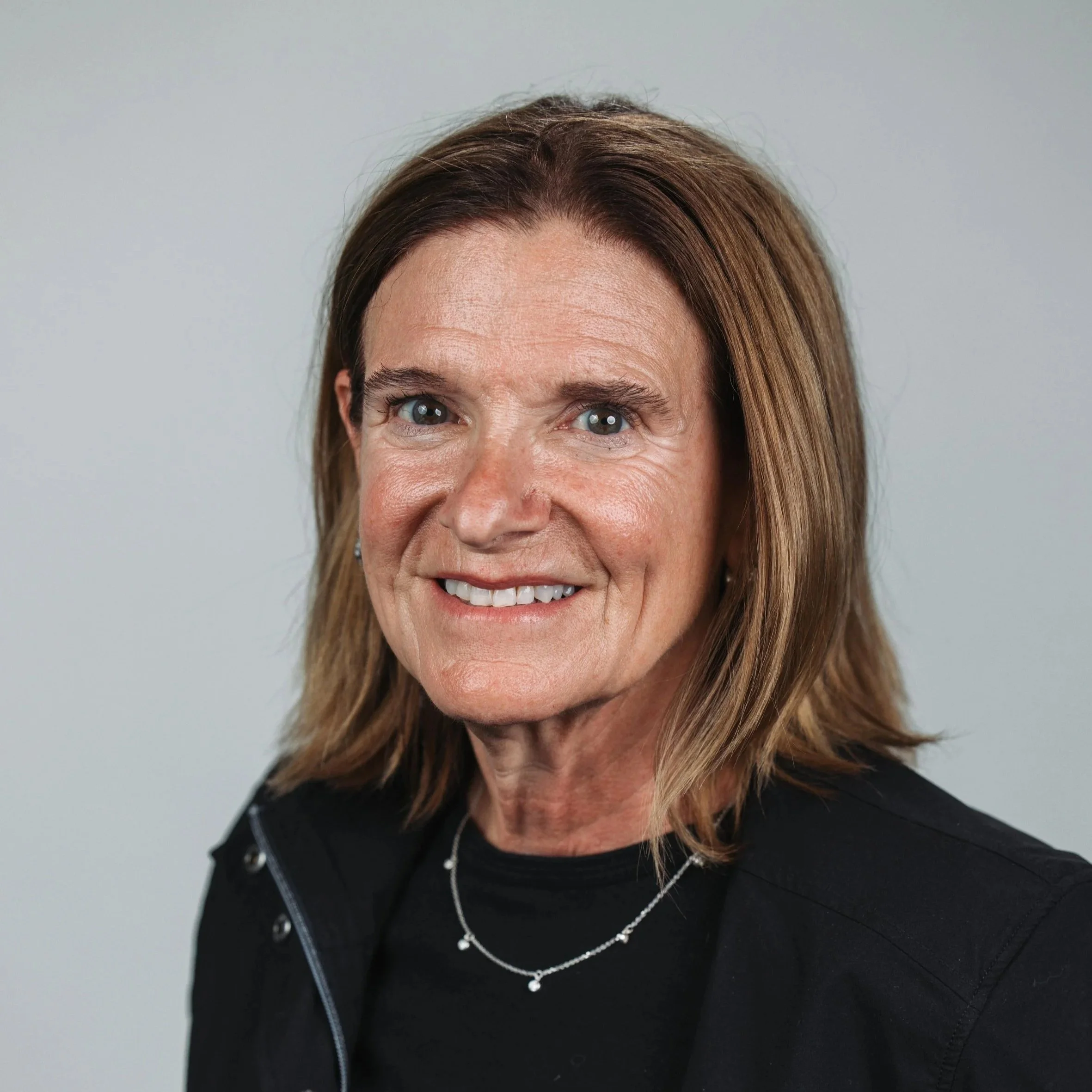 Sam Albano, board member of Charis Pregnancy Clinic, with long brown hair wearing a black shirt, standing against a plain light gray background.