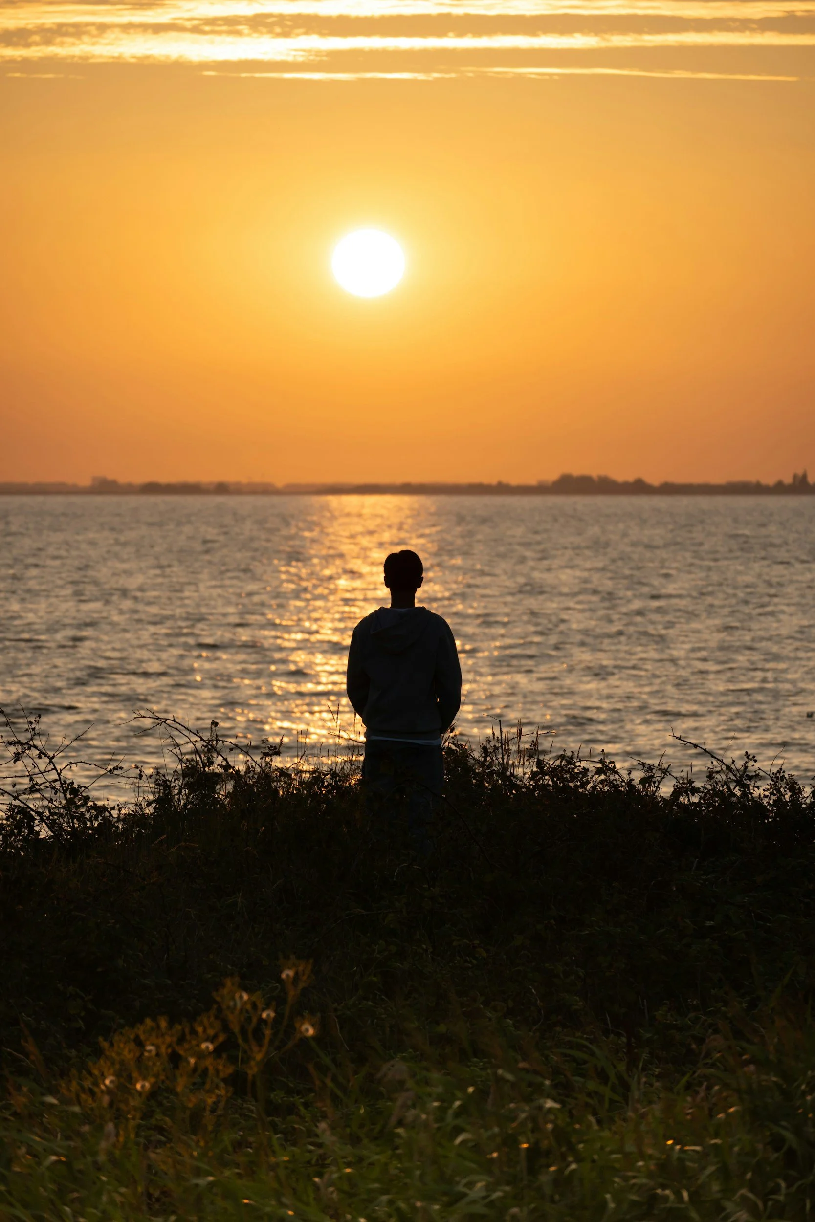 Person standing on the beach watching the sunset over the water with orange sky and reflections.