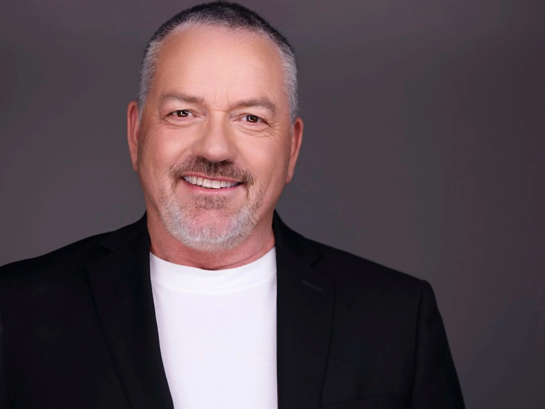 A middle-aged man with gray hair and a beard, smiling, wearing a black suit and white shirt, against a gray background.