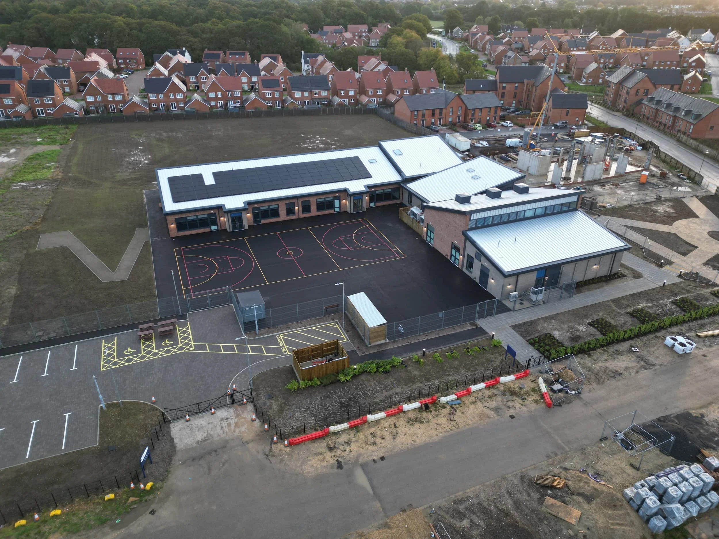 Aerial view of a newly constructed school building with a basketball court in the foreground, located next to a suburban residential area with numerous brick houses. Based in Dorset, Somerset, Wiltshire, Hampshire, West Sussex, East Sussex, Surrey, B