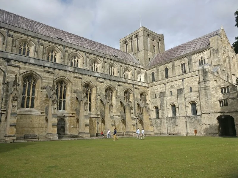 Exterior view of historic Winchester, Hampshire cathedral with tall, narrow stained glass windows and flying buttresses, surrounded by a grassy courtyard with a few people walking. Overcast sky.