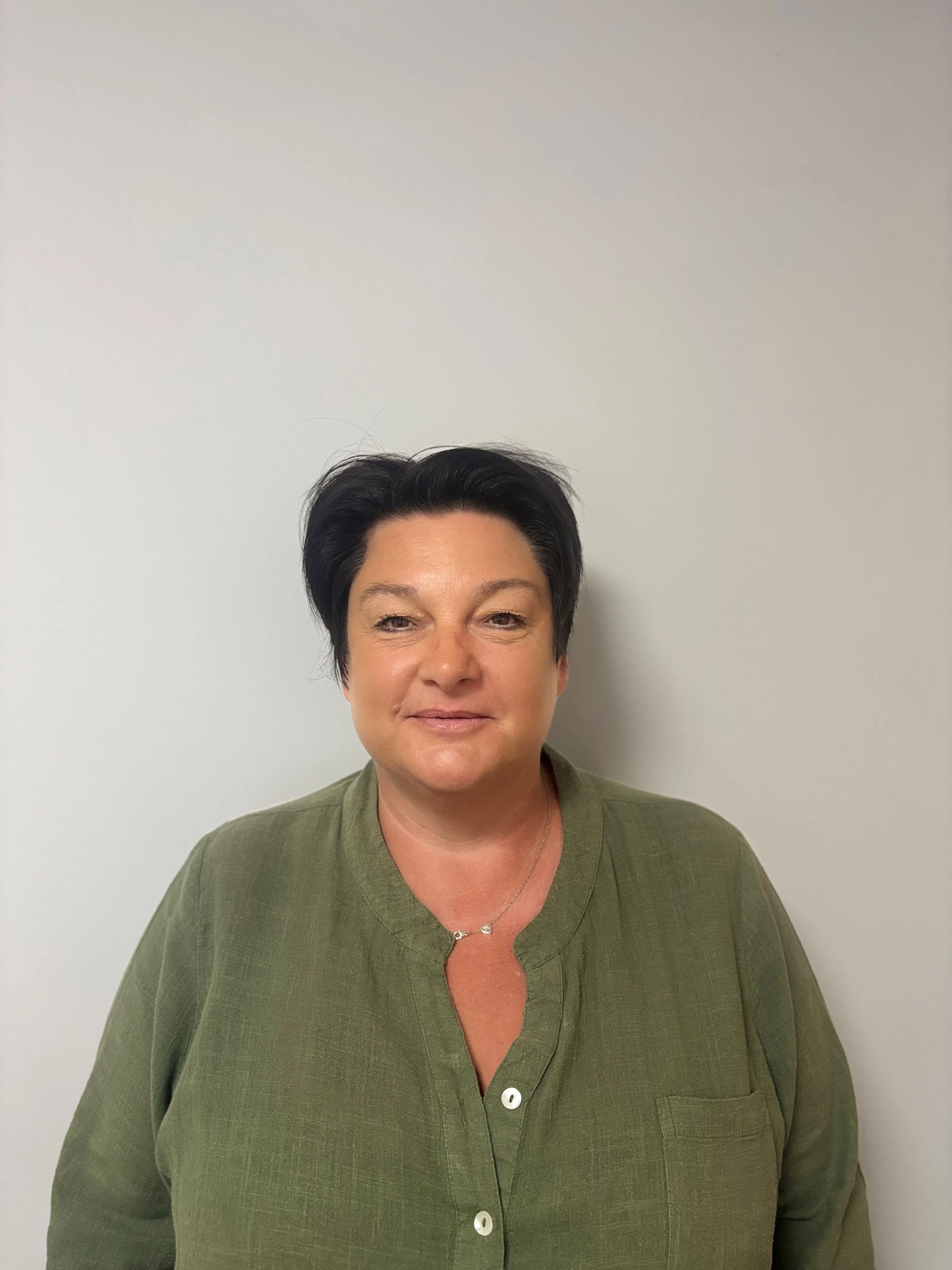 A woman with short dark hair, wearing a green top and a delicate necklace, standing against a plain light-colored wall.