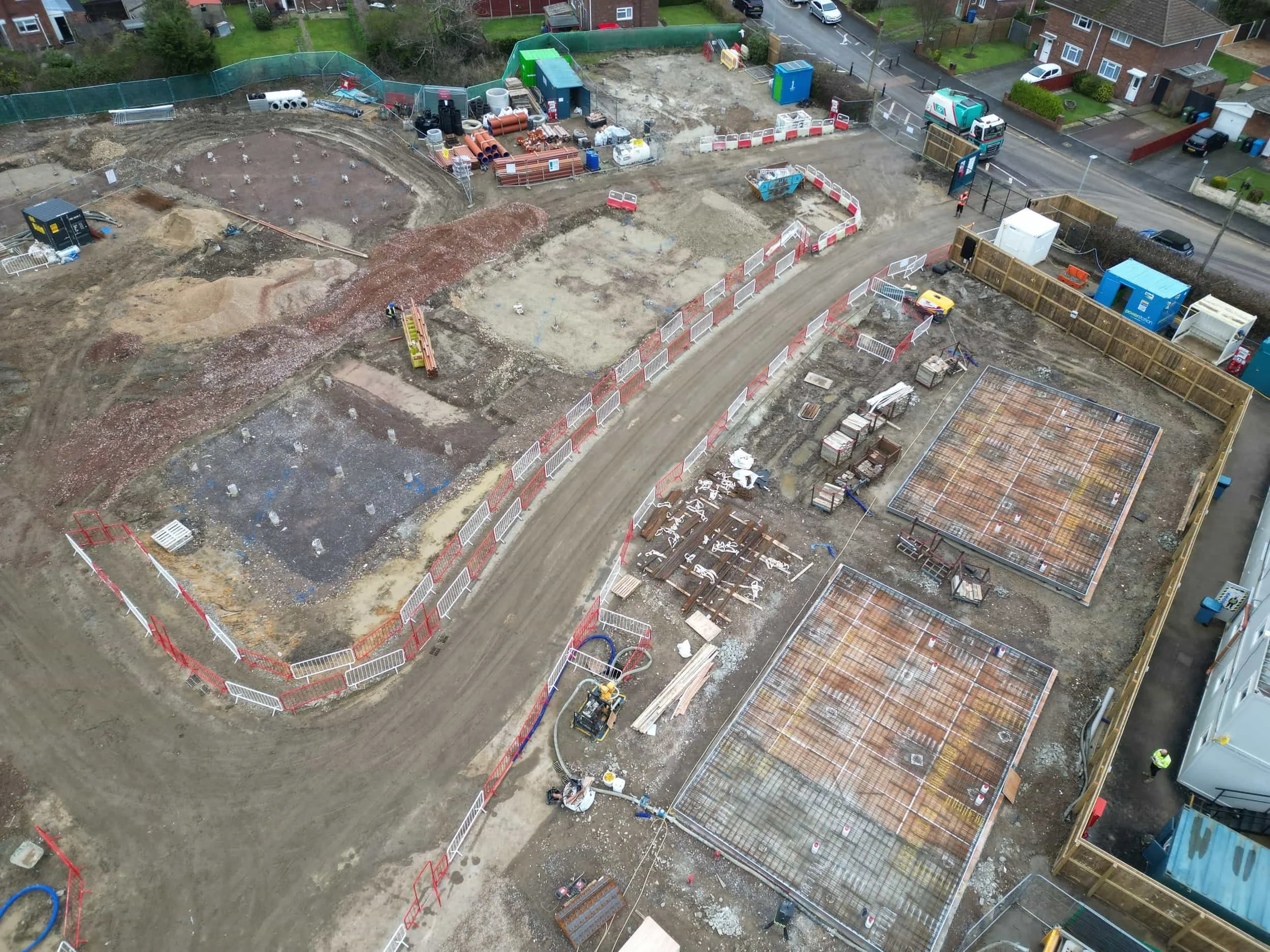 Aerial view of a construction site with areas prepared for foundations, construction materials, equipment, and fenced perimeter, adjacent to residential houses.