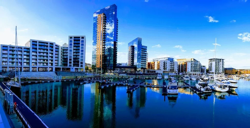 Southampton, Hampshire marina with yachts and sailboats docked, modern high-rise buildings reflecting in the water, under a clear blue sky with a few clouds.