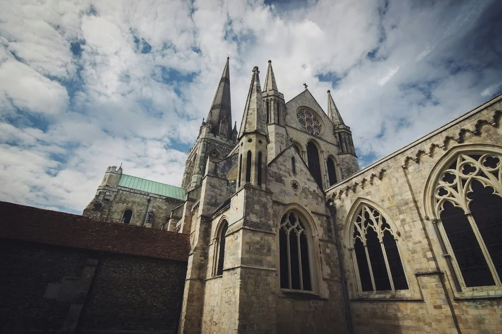 Low-angle view of Chichester, West Sussex cathedral with tall spires and ornate windows under a partly cloudy sky.