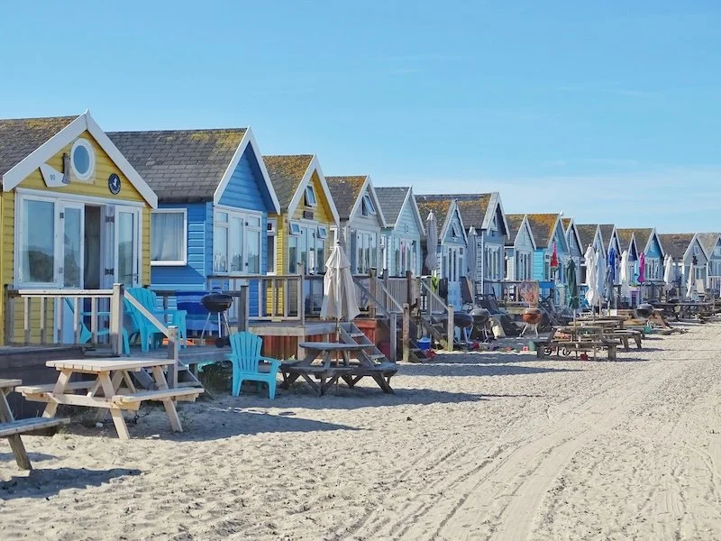 Colorful beach houses in Bournemouth, Dorchester, lined up on sandy shore with chairs, tables, umbrellas, and a clear blue sky.