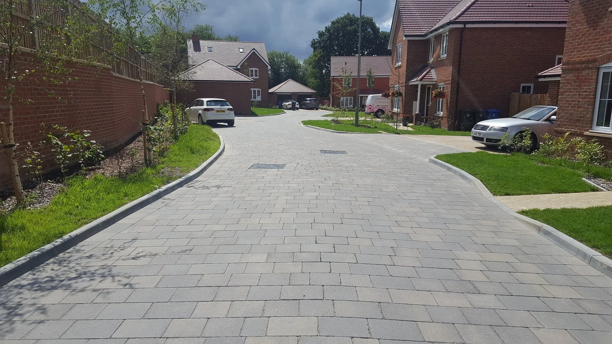 Residential neighborhood street with paved driveway, brick houses, parked cars, green lawns, and trees.