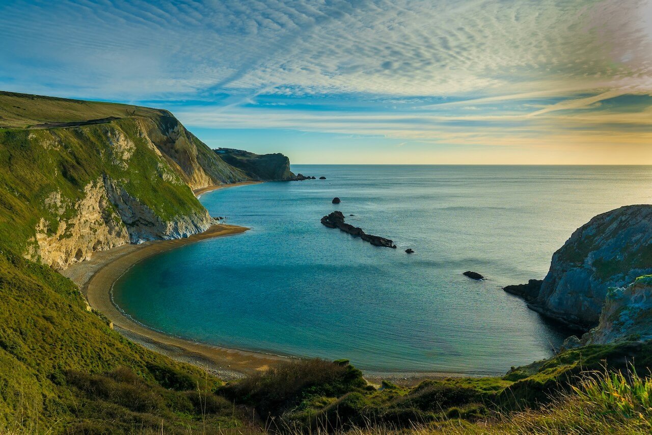 A scenic view of Bridport, Dorset, a crescent-shaped beach surrounded by high cliffs with green vegetation, calm blue water, and a partly cloudy sky at sunset or sunrise.