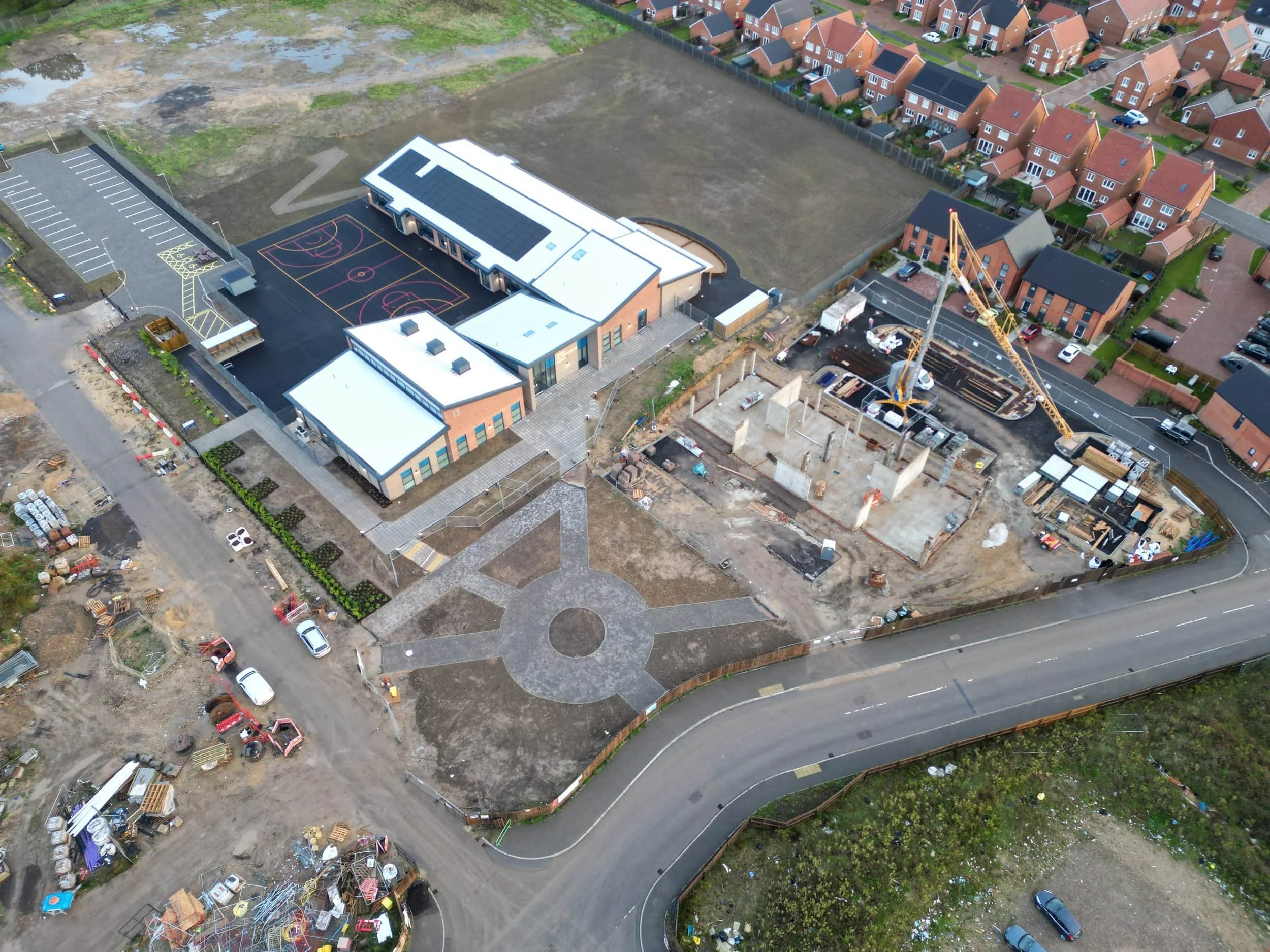 Aerial view of a developing area with a newly constructed building featuring a sports court, adjacent construction site with heavy machinery, and nearby residential homes. Based in Dorset, Somerset, Wiltshire, Hampshire, West Sussex, East Sussex, Sur