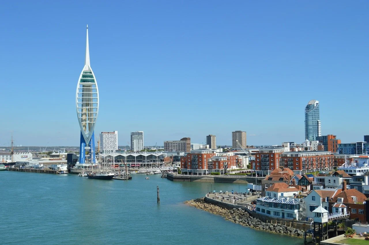 Portsmouth, Hampshire skyline with sail-shaped skyscraper next to harbor, including boats and waterfront buildings under a clear blue sky.