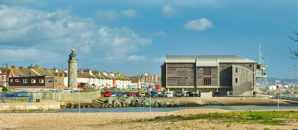 View of a Worthing, West Sussex with modern building, lighthouse, and row of houses with parked cars along the street. The sky is partly cloudy.